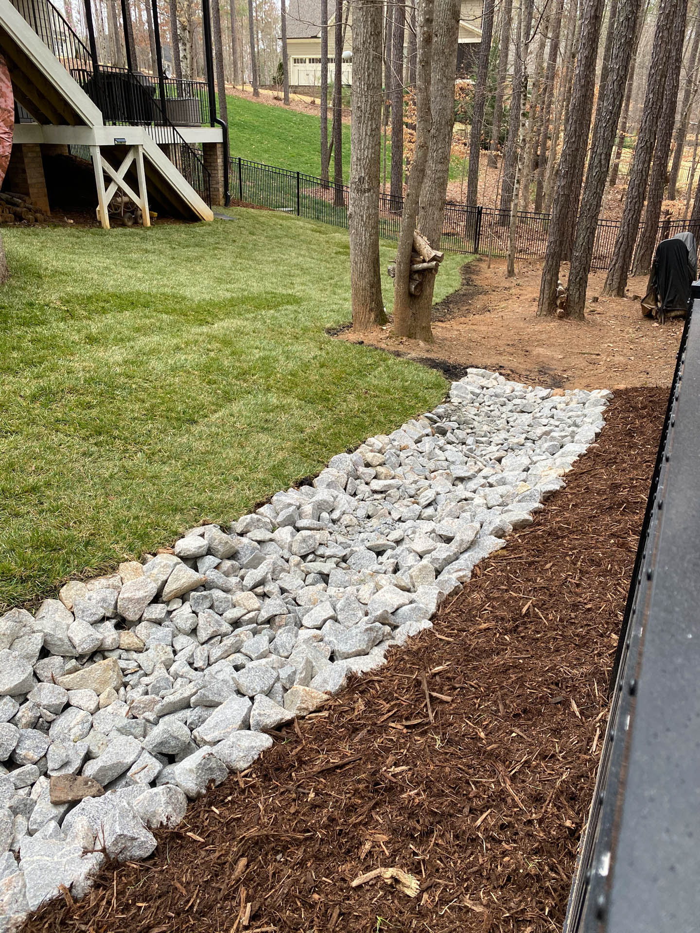 Rock-lined drainage channel next to a mulched area, separating a lawn and a tree-lined area.