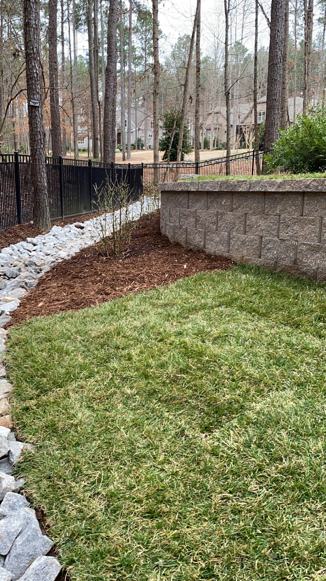 A landscaped yard with a grassy area, mulch, rocks, and a stone retaining wall. Trees are in the background.