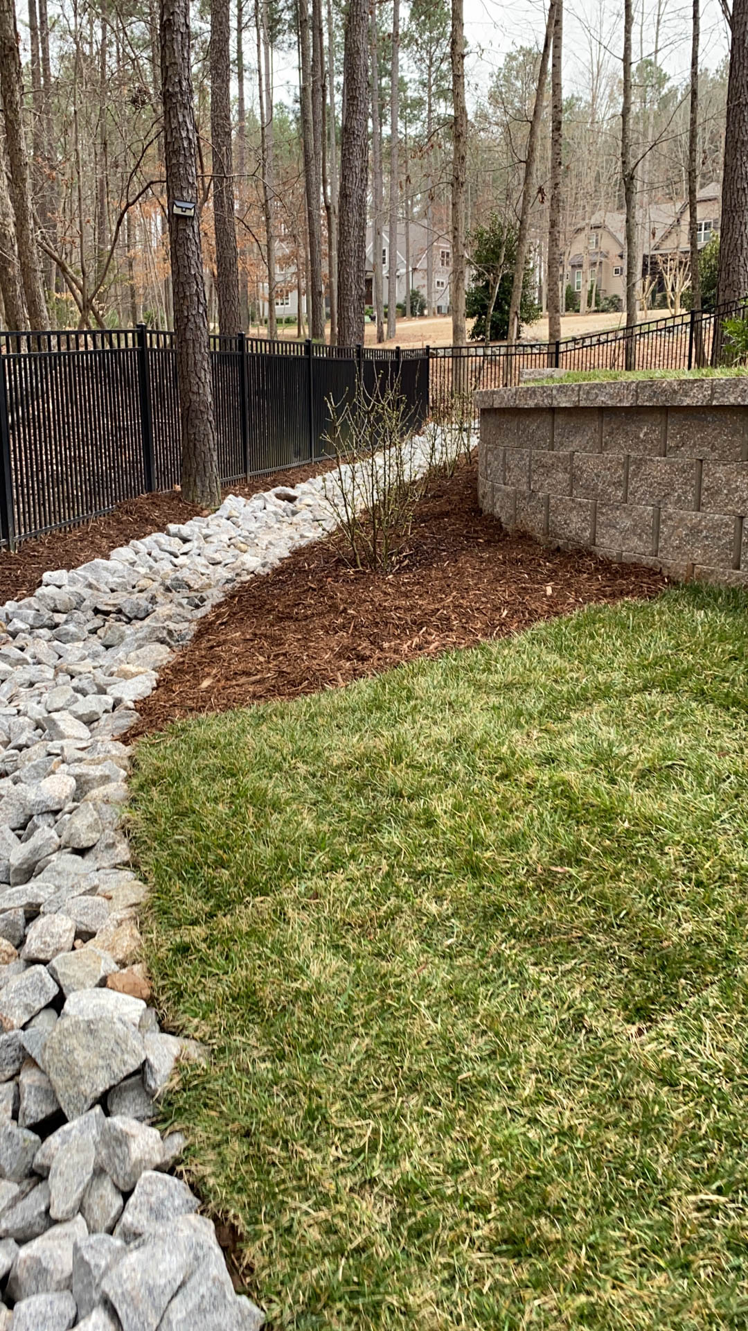Gravel-lined drainage channel next to a green lawn, edged with mulch, trees, and a retaining wall.