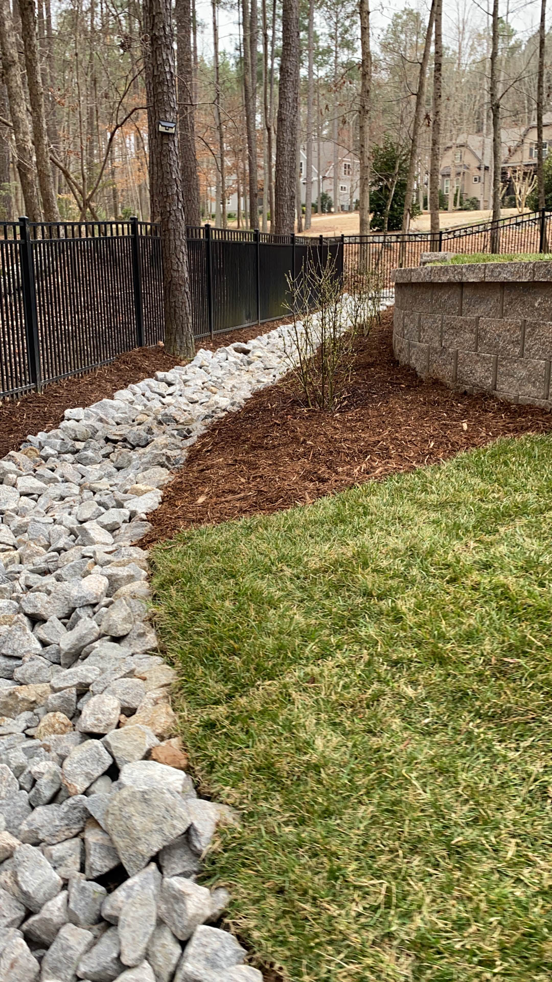 A rock-lined drainage channel with grass and mulch next to a fence and a retaining wall.