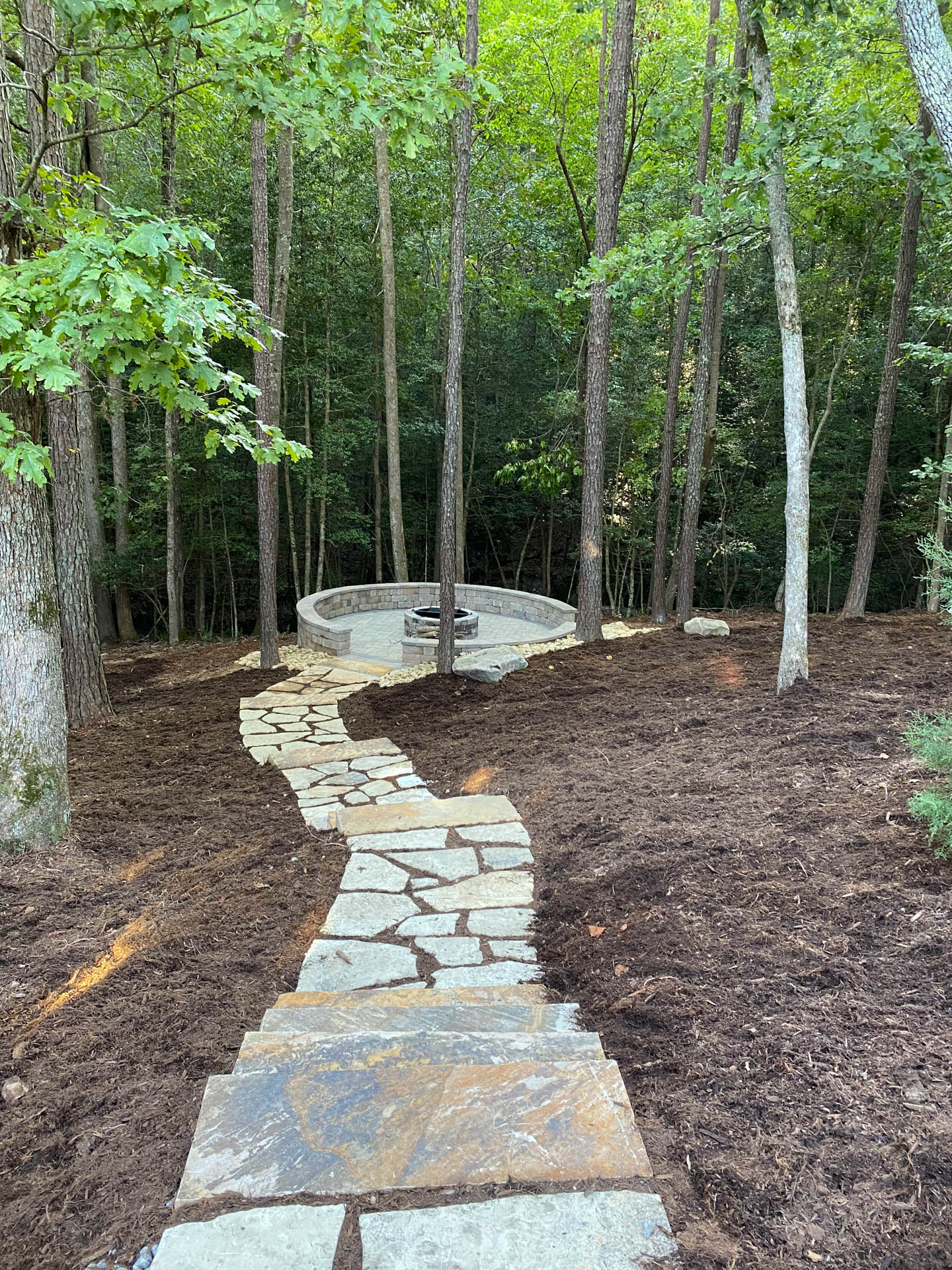 Stone path leading to a fire pit in a wooded area. The ground is covered in mulch.