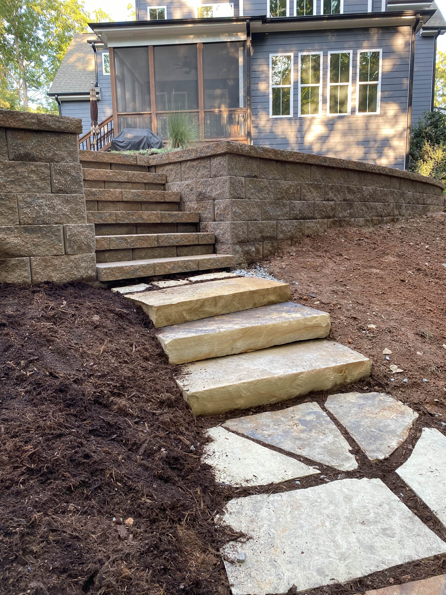Stone steps leading up a slope, with a retaining wall and house in the background. Mulch and landscaping surround.