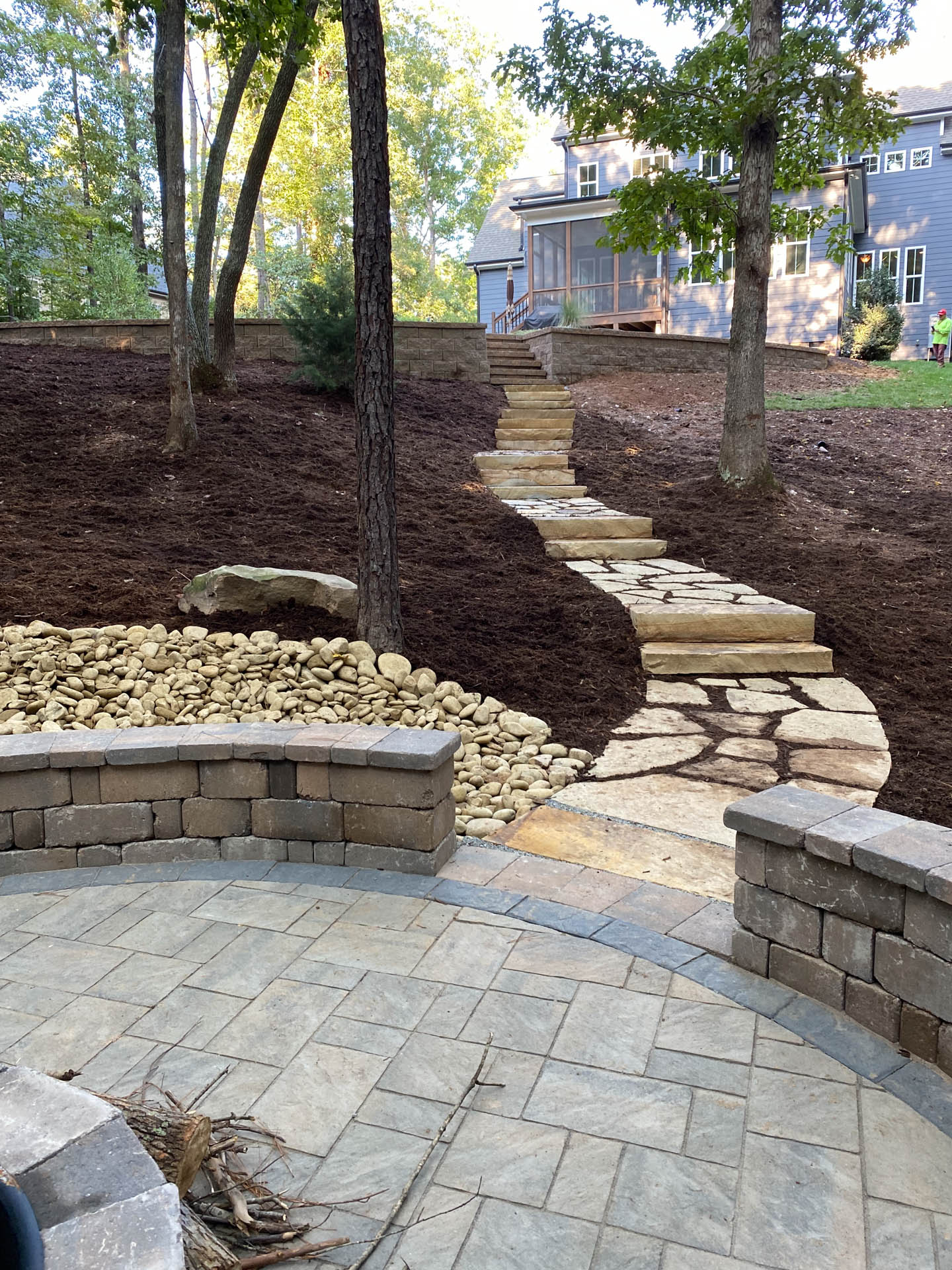 Stone patio with steps leading up a mulched hill to a house.
