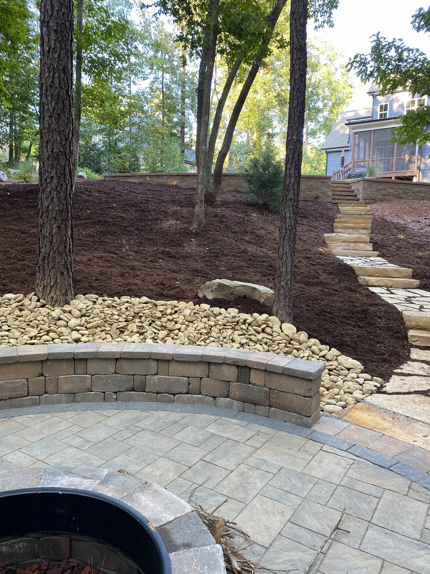 Stone patio with fire pit, retaining walls, steps, and trees on a mulched hillside.