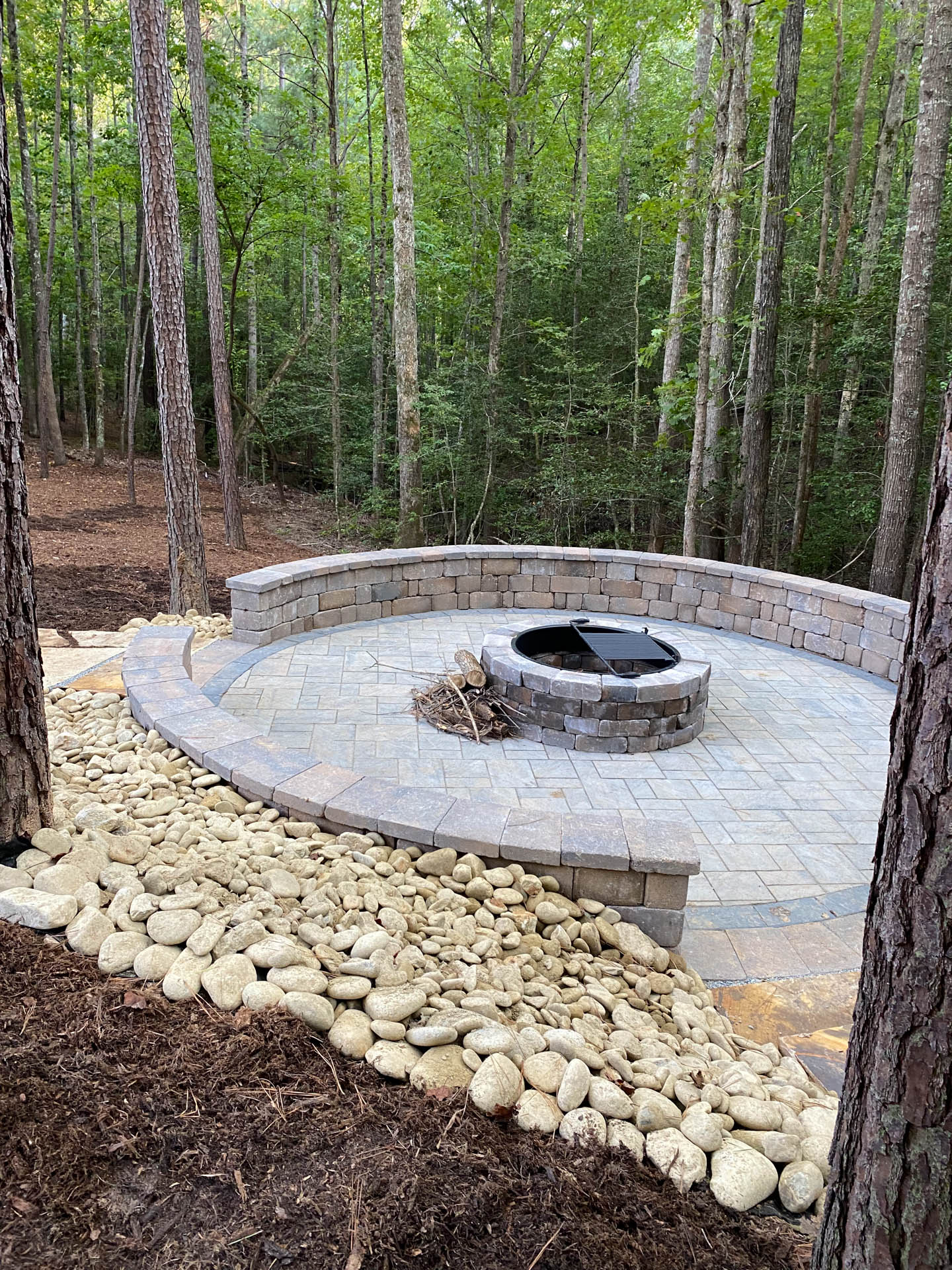 Circular stone patio with fire pit, surrounded by a low stone wall, wooded backdrop.