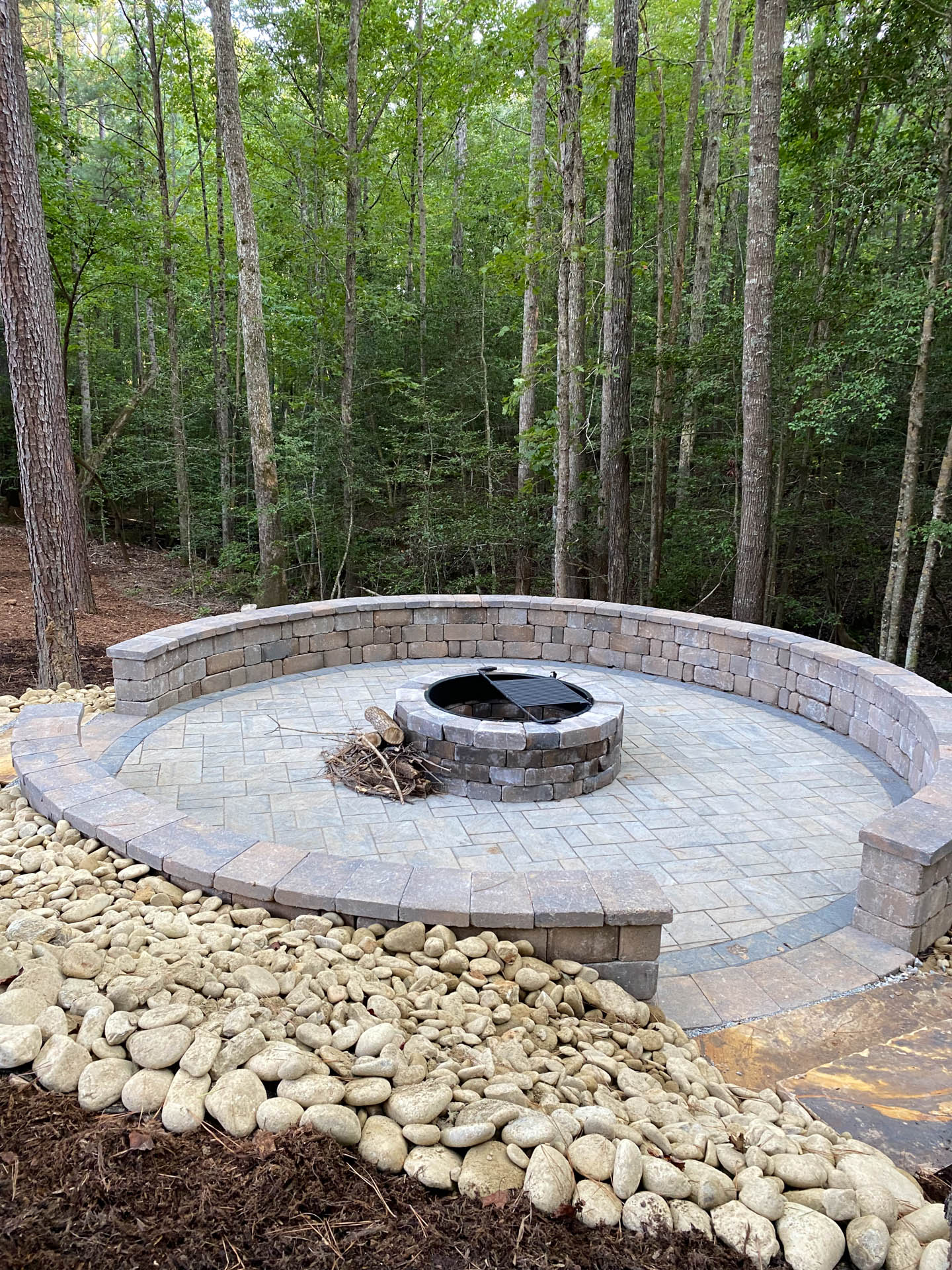 Circular stone fire pit area in a forest setting, surrounded by a stone wall and landscaping with rocks.
