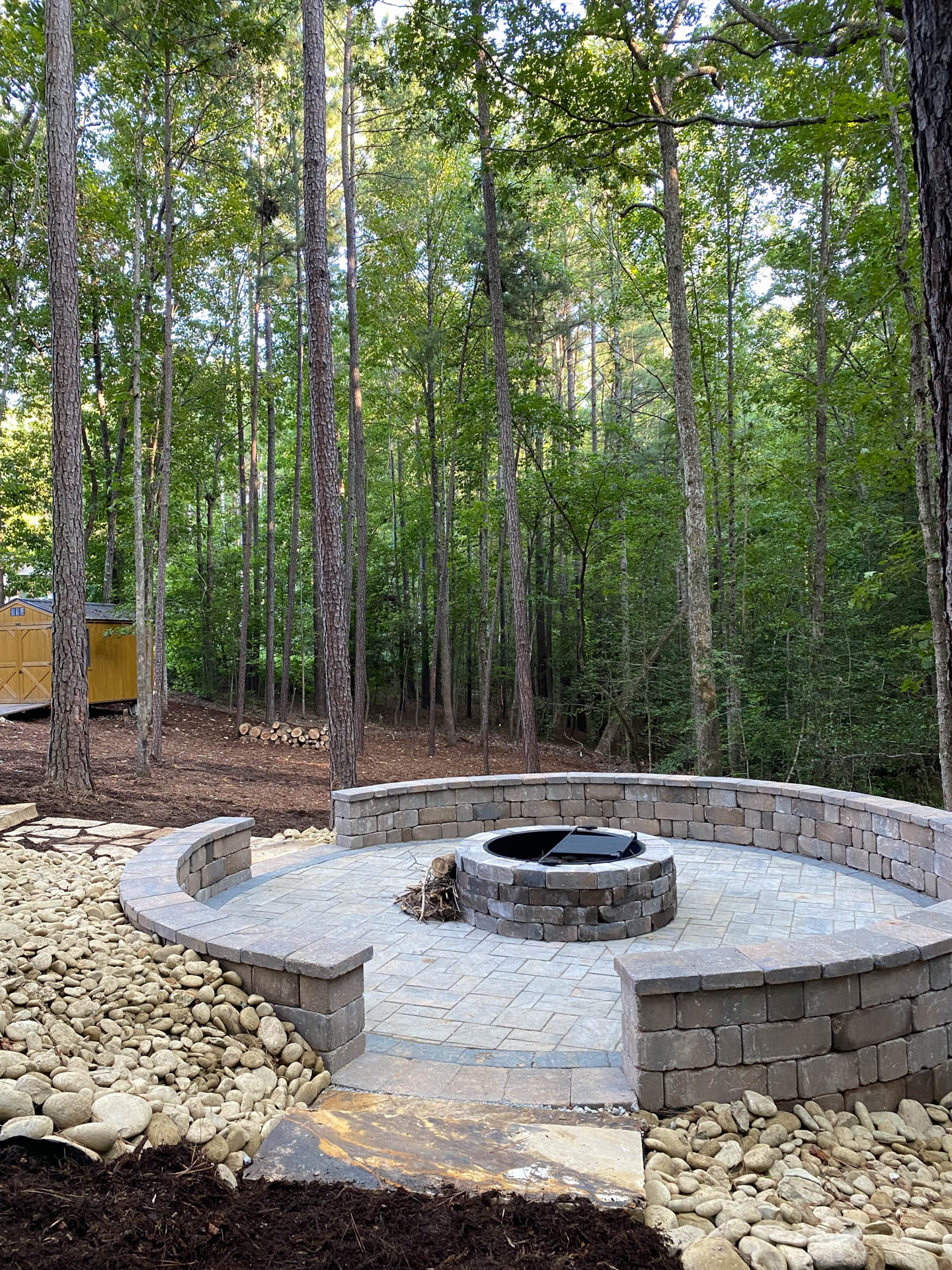 Stone fire pit in a wooded setting. Paved area with surrounding curved stone wall.