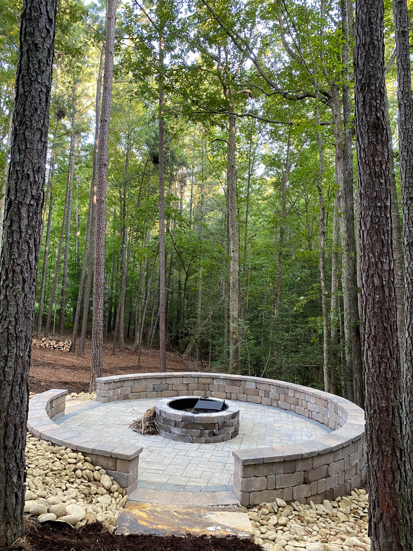 Circular stone fire pit in a forest, surrounded by trees and a retaining wall.