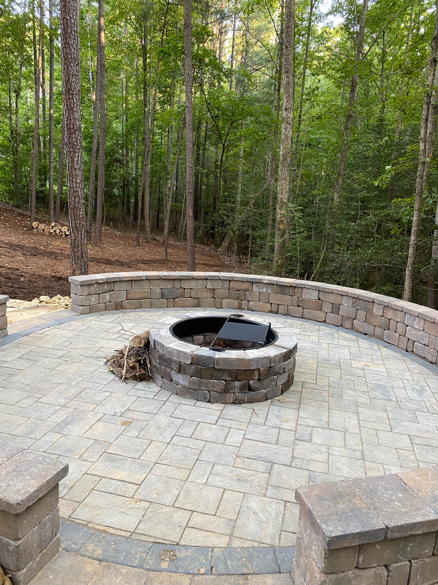 Circular stone patio with fire pit, surrounded by a low wall. Wooded background.