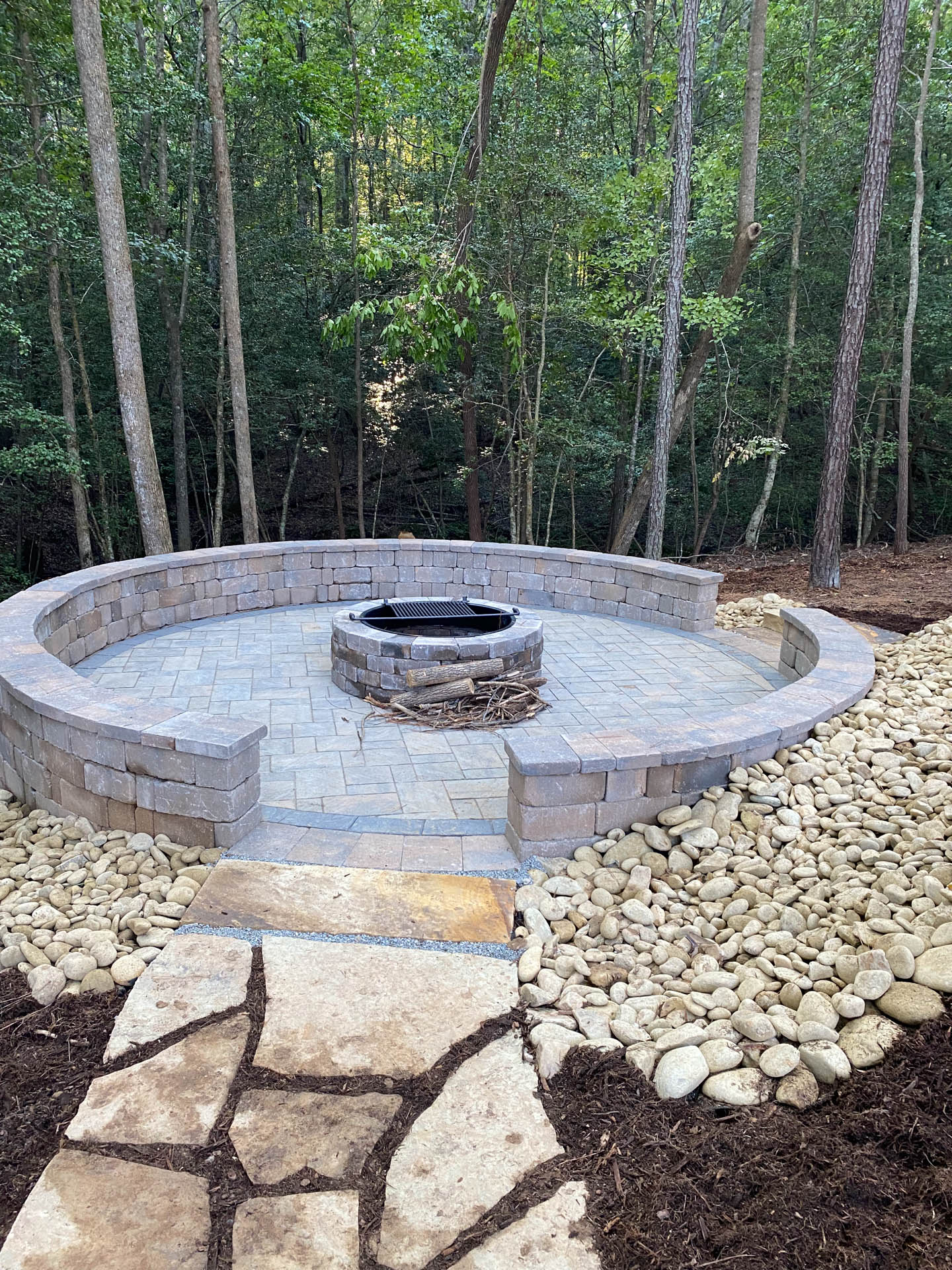 Stone fire pit in a circular patio, pathway leading to it. Brown stones and mulch surround.