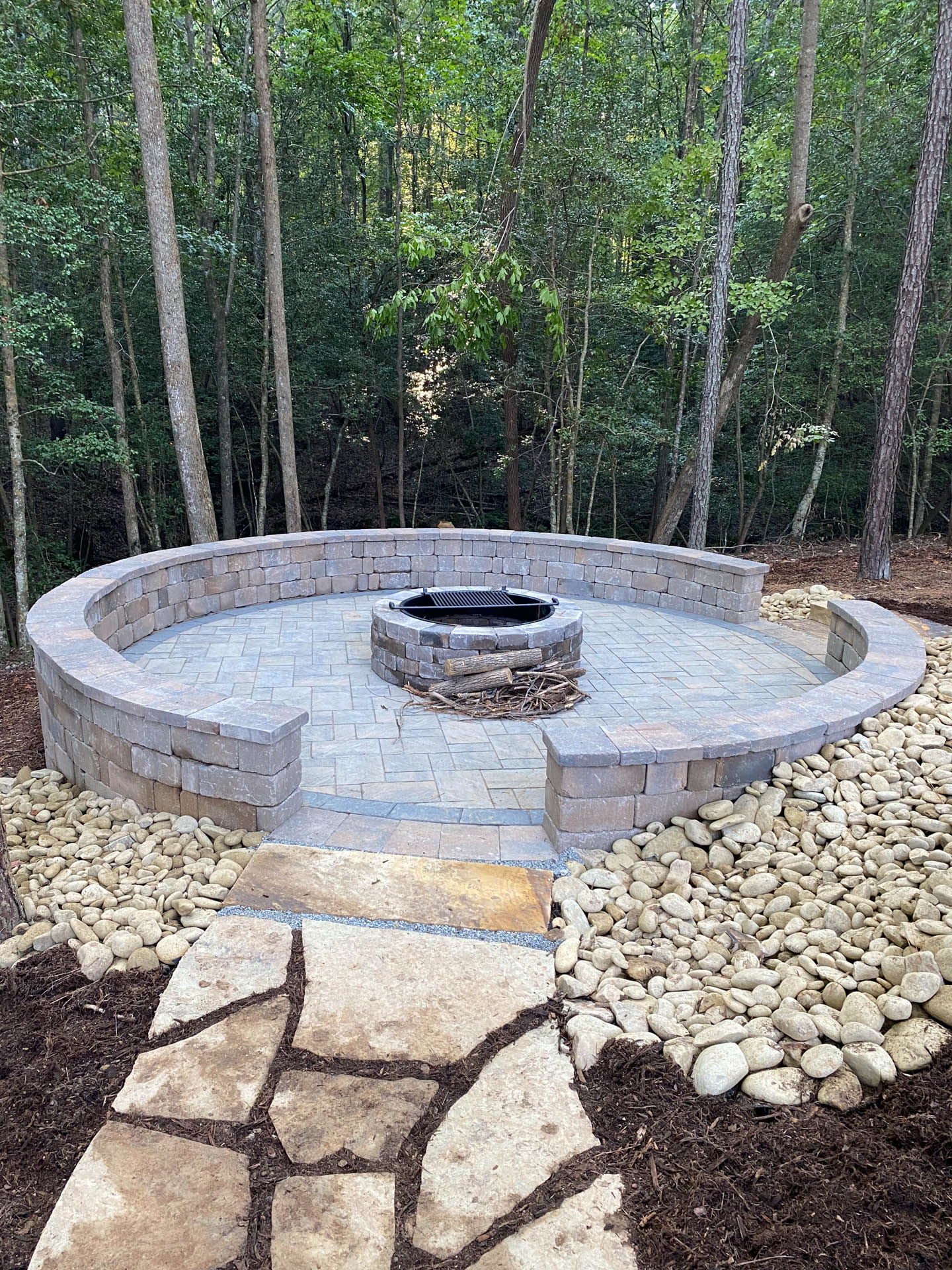 Stone fire pit with circular seating area in a wooded setting. Pathway leads to the pit.