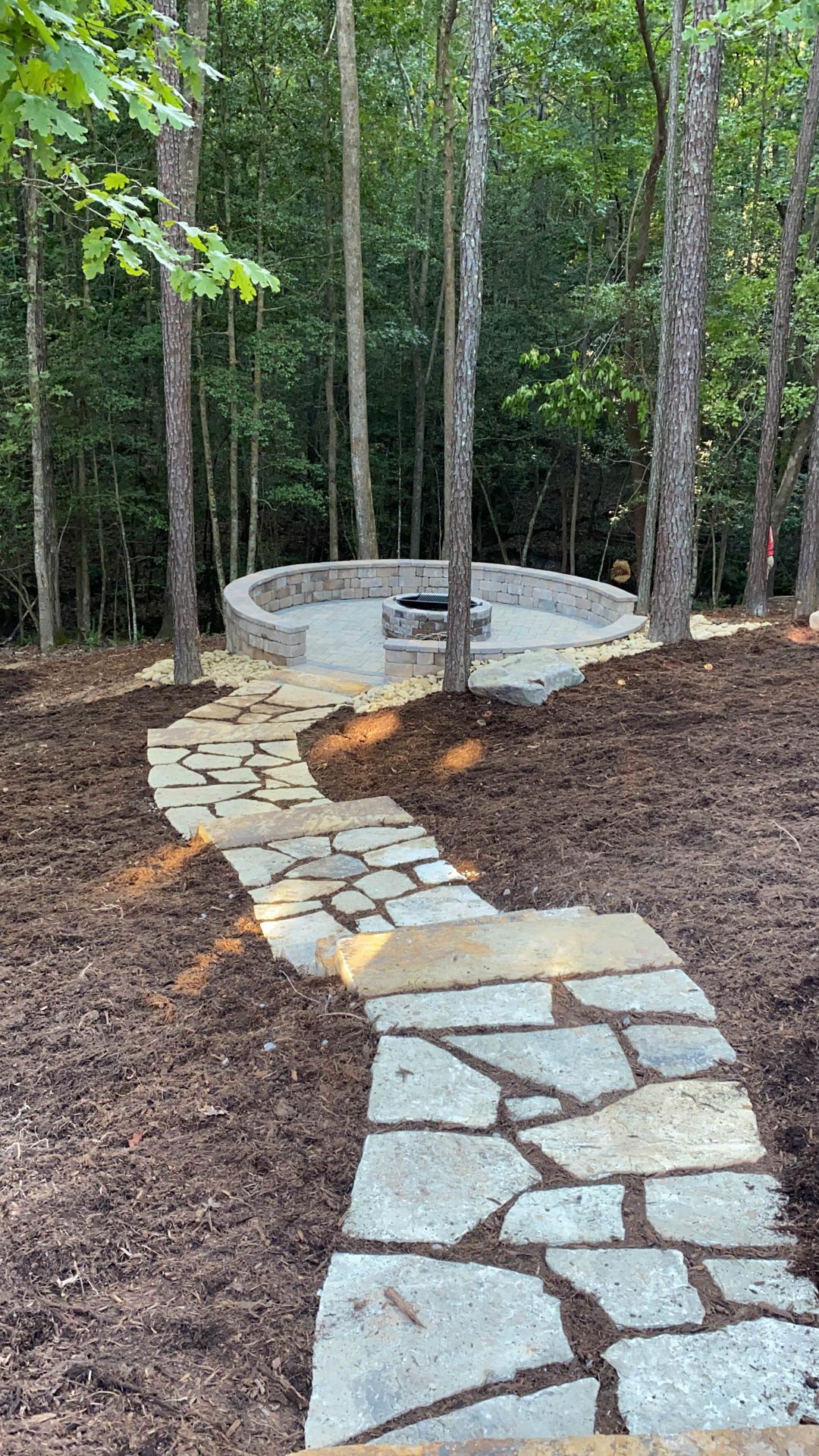 Stone pathway leads to a circular fire pit in a wooded area. Brown mulch surrounds the path.