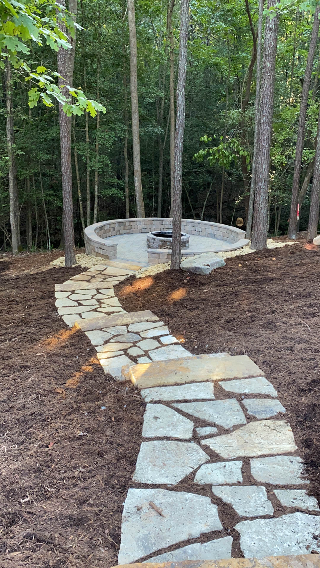 Stone pathway leading to a circular patio with a fire pit, set in a wooded area.