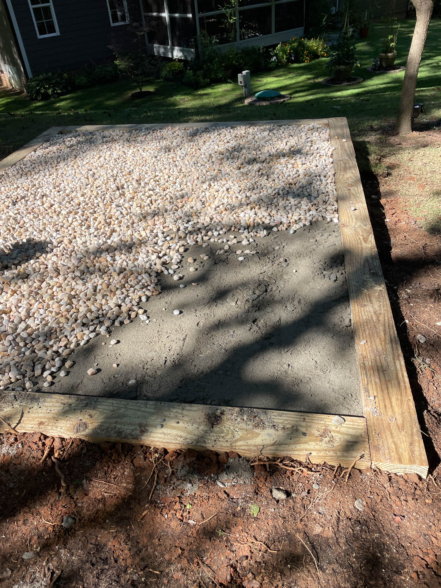 Gravel and concrete fill a wooden-framed rectangular area on a patch of dirt, likely a prepared surface for a structure.