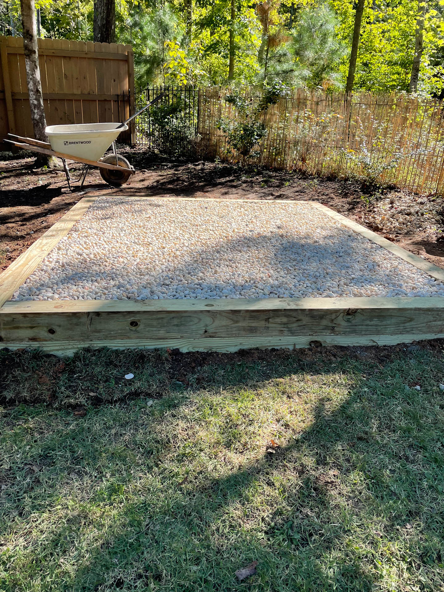 Gravel-filled square platform with wooden borders on grass. A wheelbarrow sits nearby in a yard.