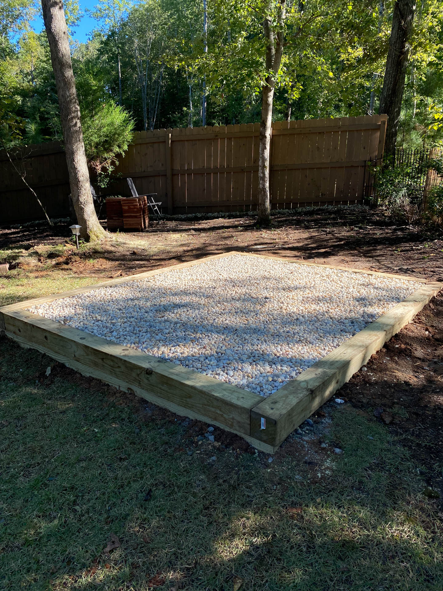 Gravel-filled rectangular base bordered by wood, surrounded by grass and trees. A wooden fence is in the background.