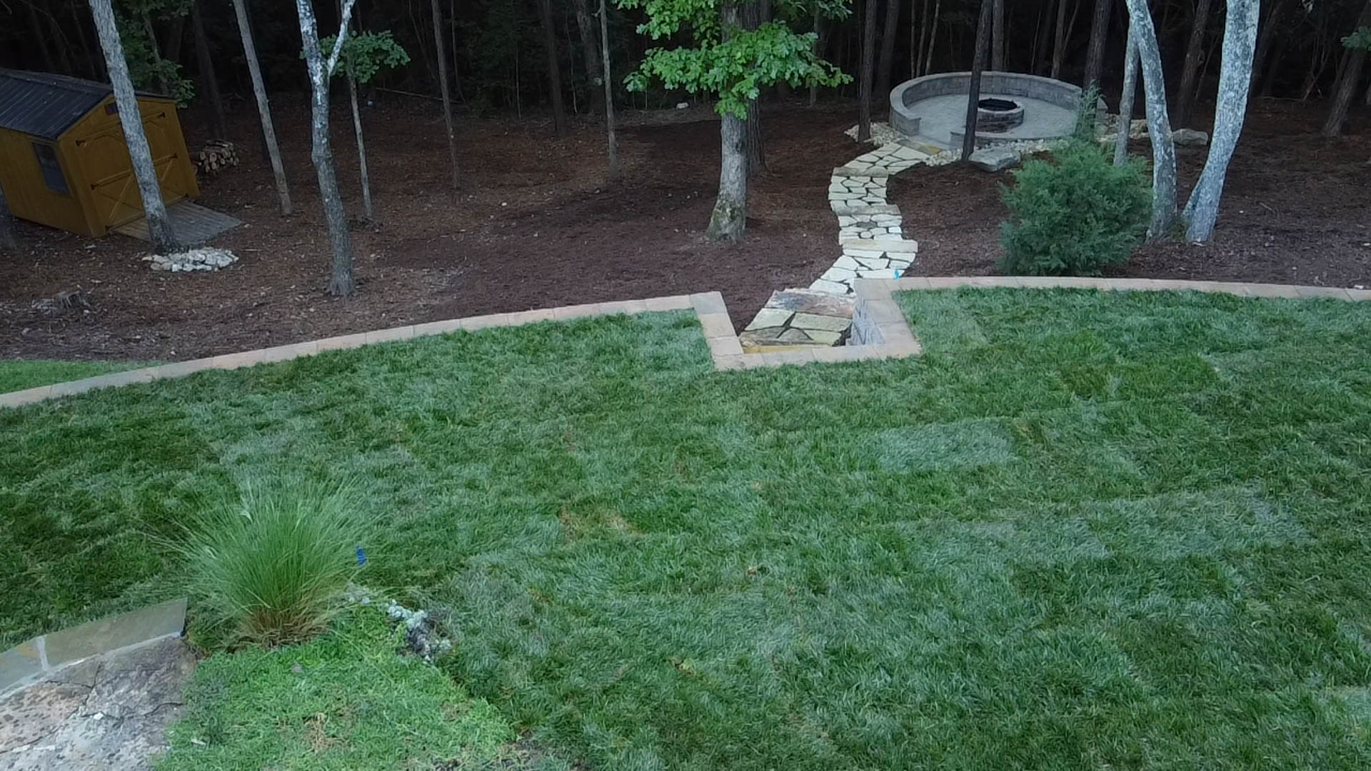 Green lawn with a stone pathway leading to a fire pit, surrounded by trees.