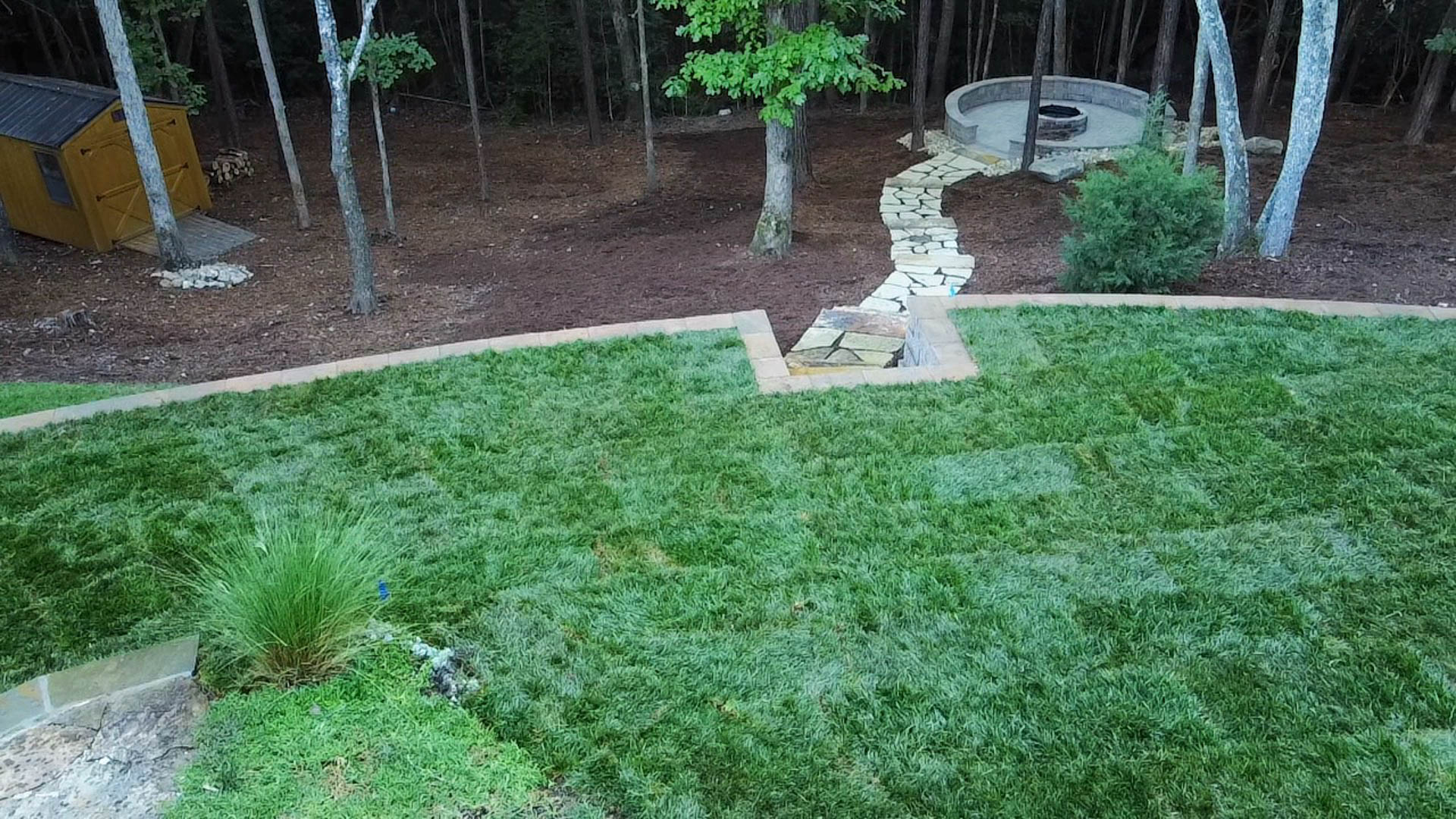 Lush green lawn leading to a stone patio with seating, fire pit, and shed in a wooded area.