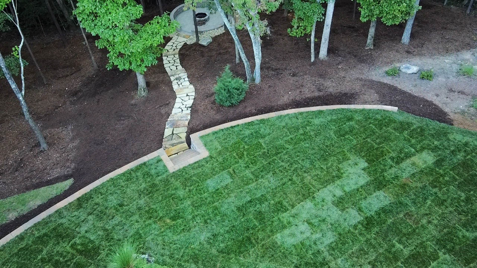 Stone path curves through trees and mulch to a small structure, beside a curved lawn.