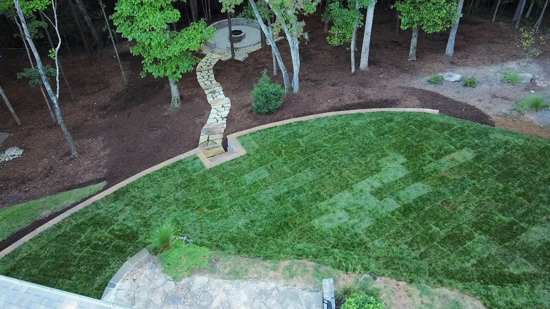 Lush green lawn borders a mulched area with a stone path leading into a wooded backdrop.