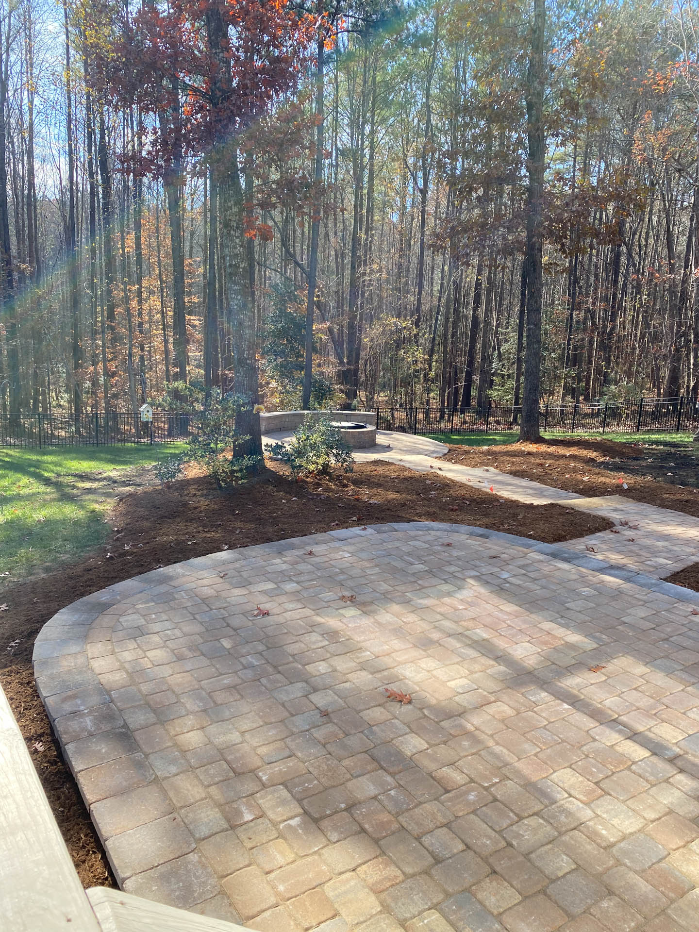Brick patio with curved edge, surrounded by mulch, and path leading to forested area.