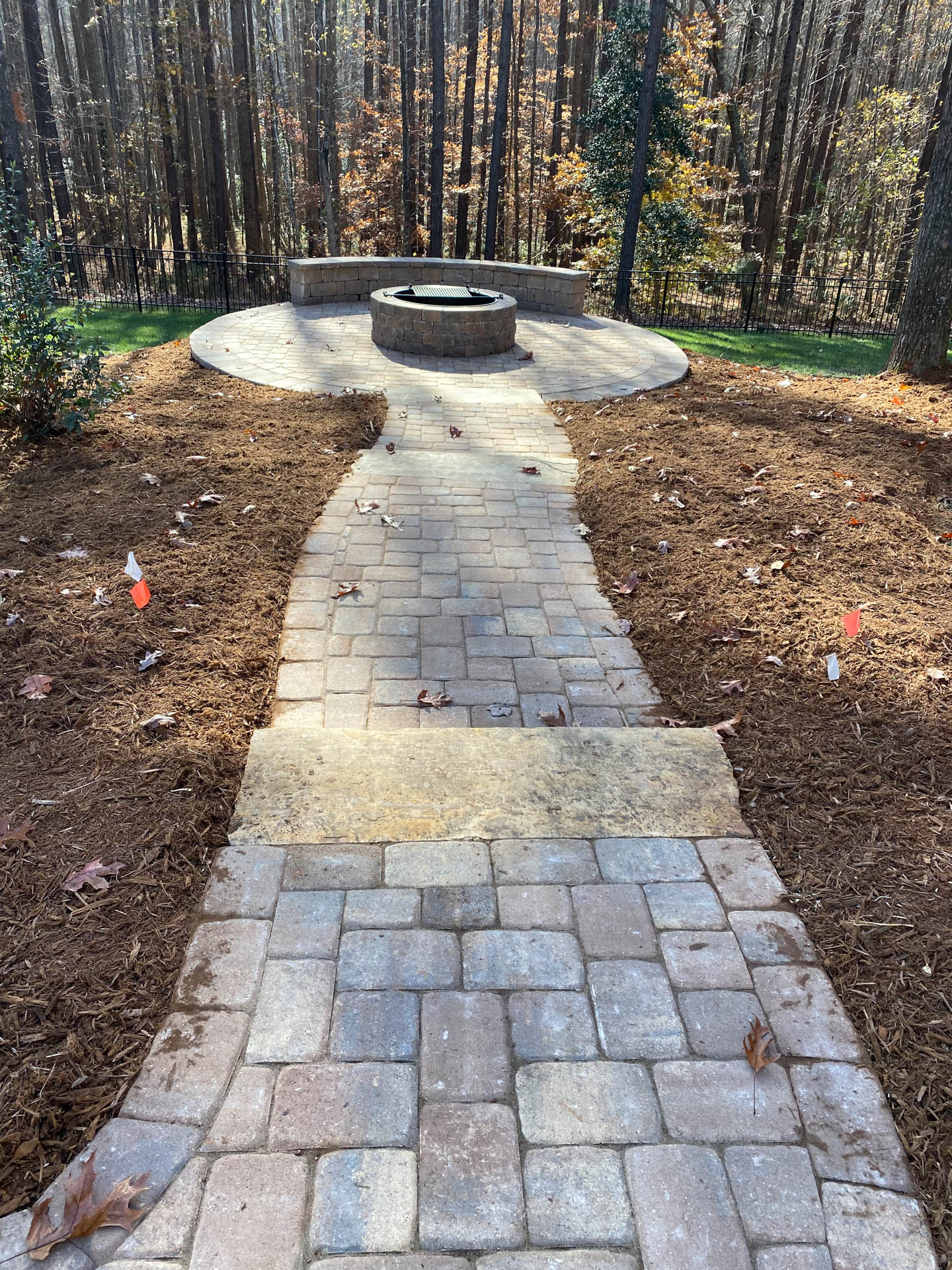 Brick pathway leads to a circular fire pit area in a wooded backyard setting, edged with mulch.