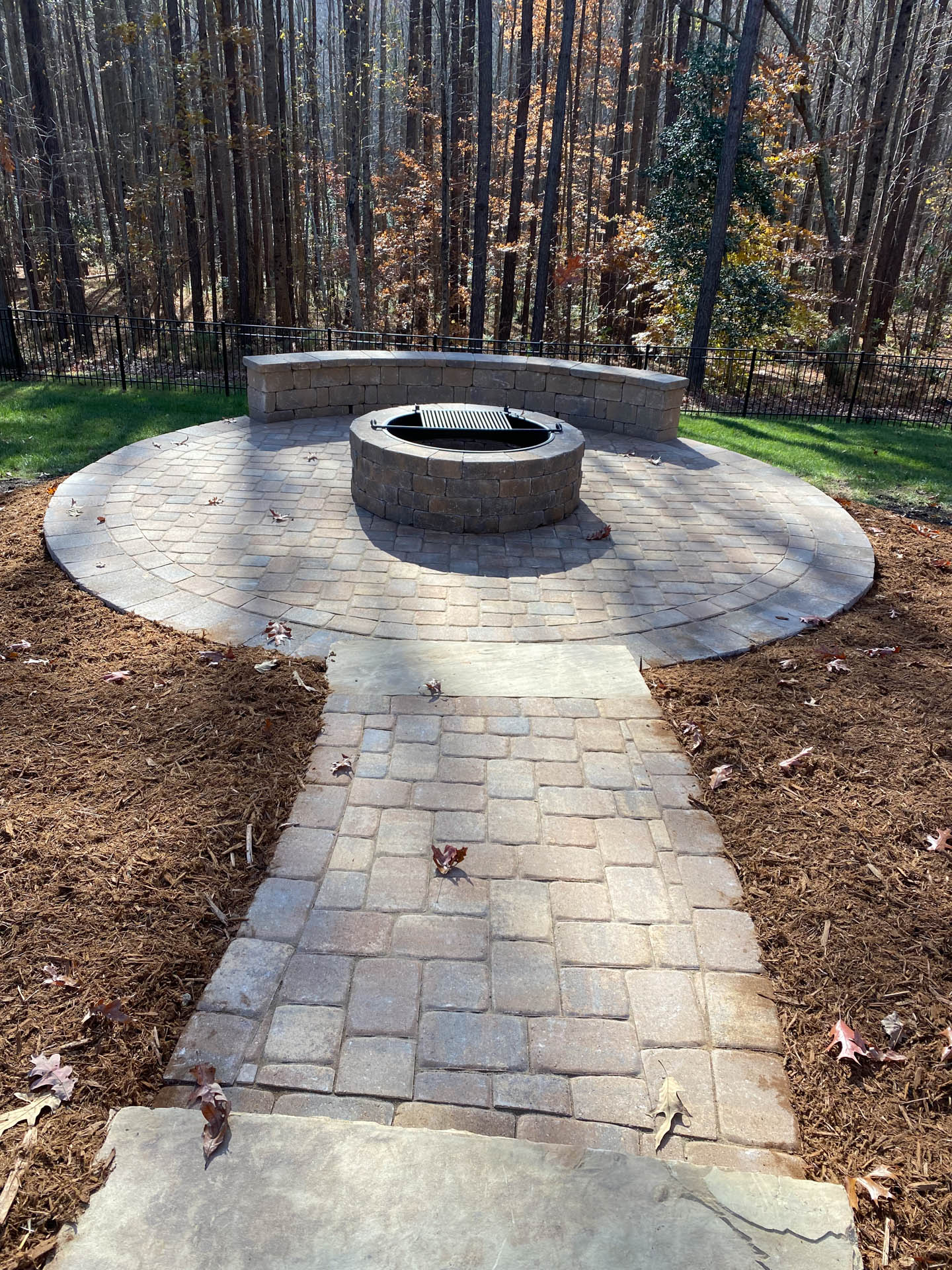 Brick patio with fire pit, bench seating, and pathway in wooded area.