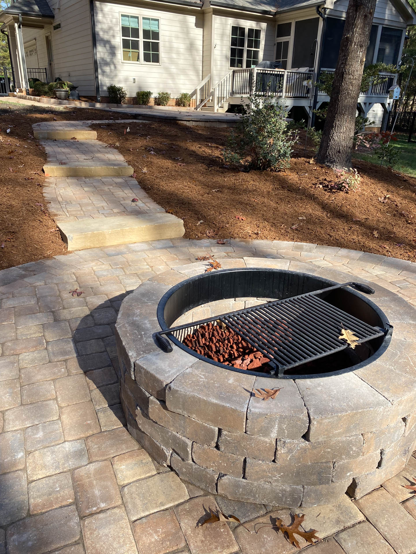 Circular brick fire pit with grill, surrounded by a paved patio and mulch. House and pathway are in the background.