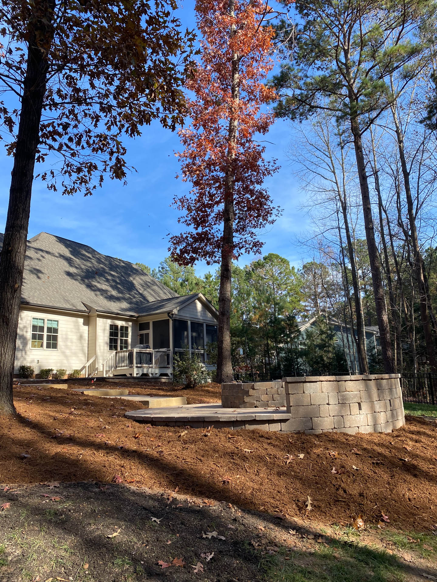 A backyard scene featuring a house, tree, and retaining wall. The tree has red leaves, and the ground is covered in mulch.