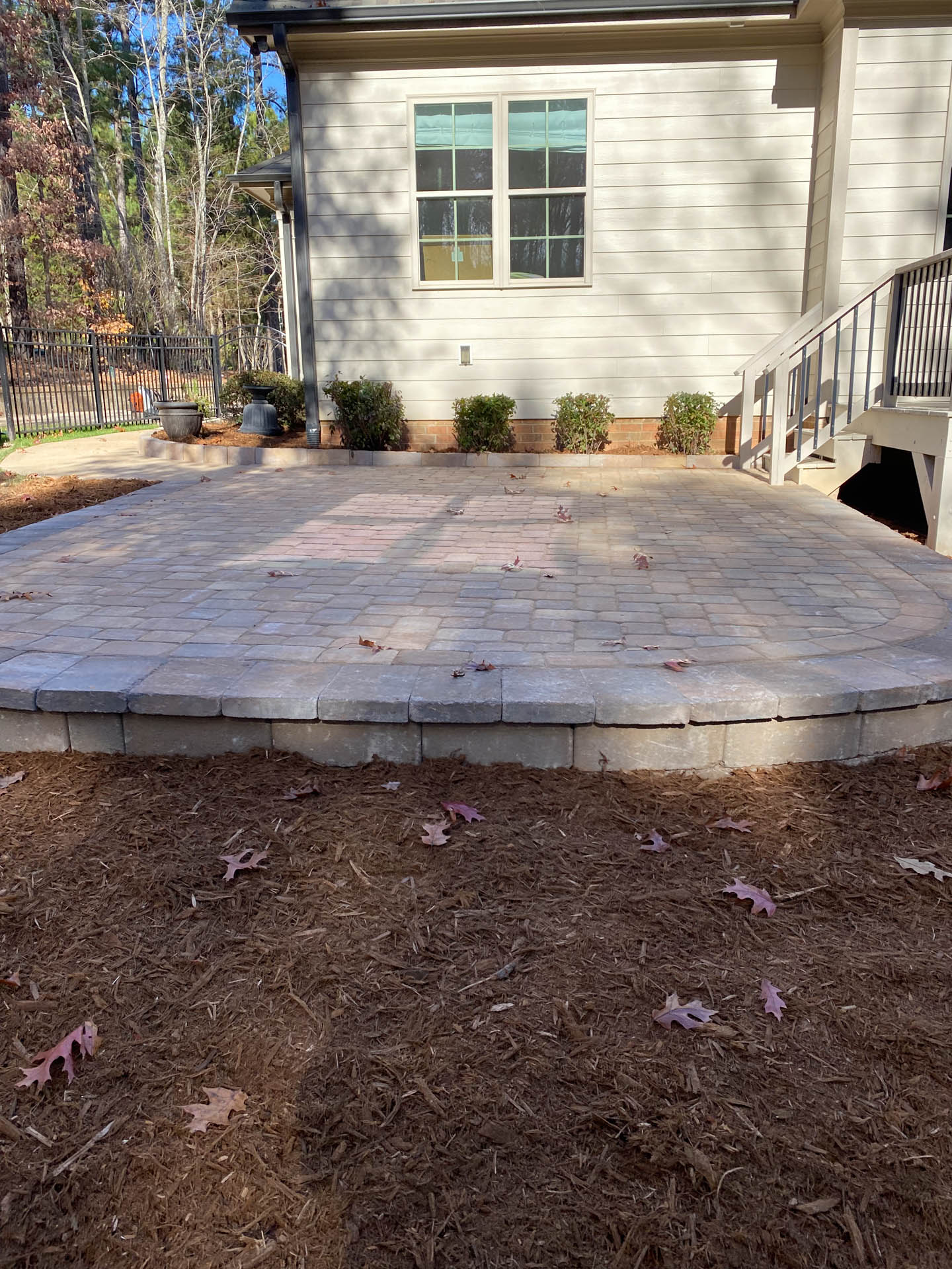 A brick patio with a curved edge, in front of a house. Mulch and small bushes are present.