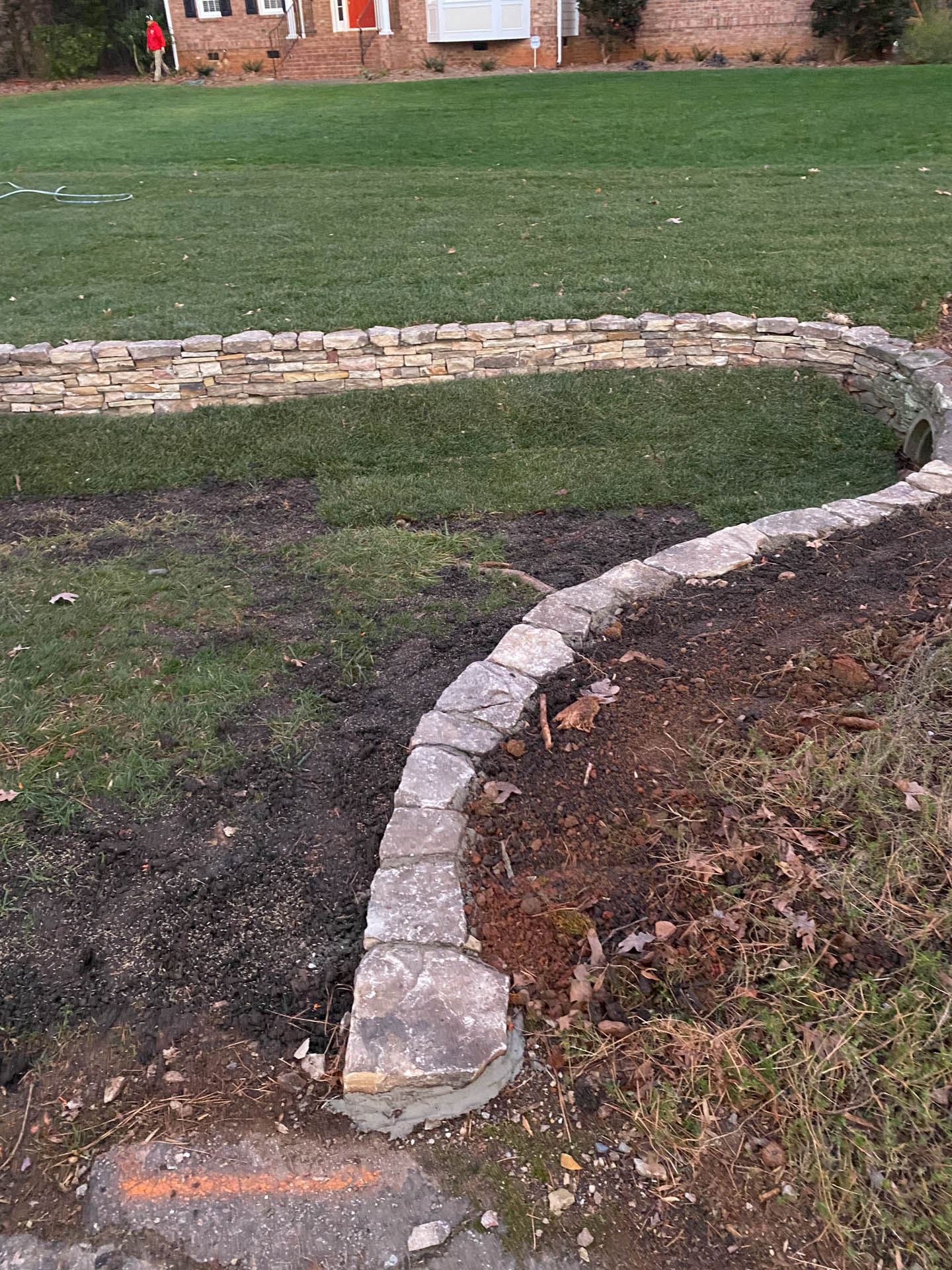 Stone retaining wall curves through a yard; person in red jacket visible in the background.