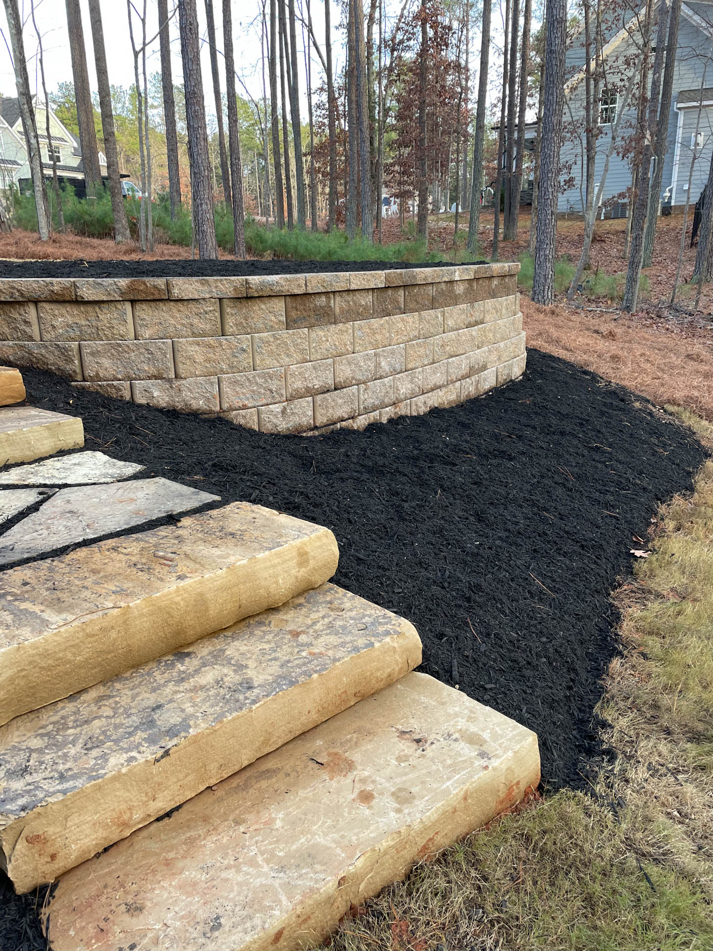 Stone steps lead to a retaining wall in a yard, covered with black mulch, wooded background.