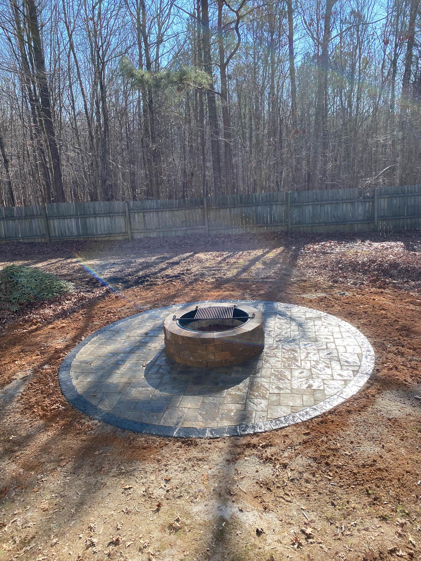 Fire pit on a circular stone patio in a backyard with trees and a fence.