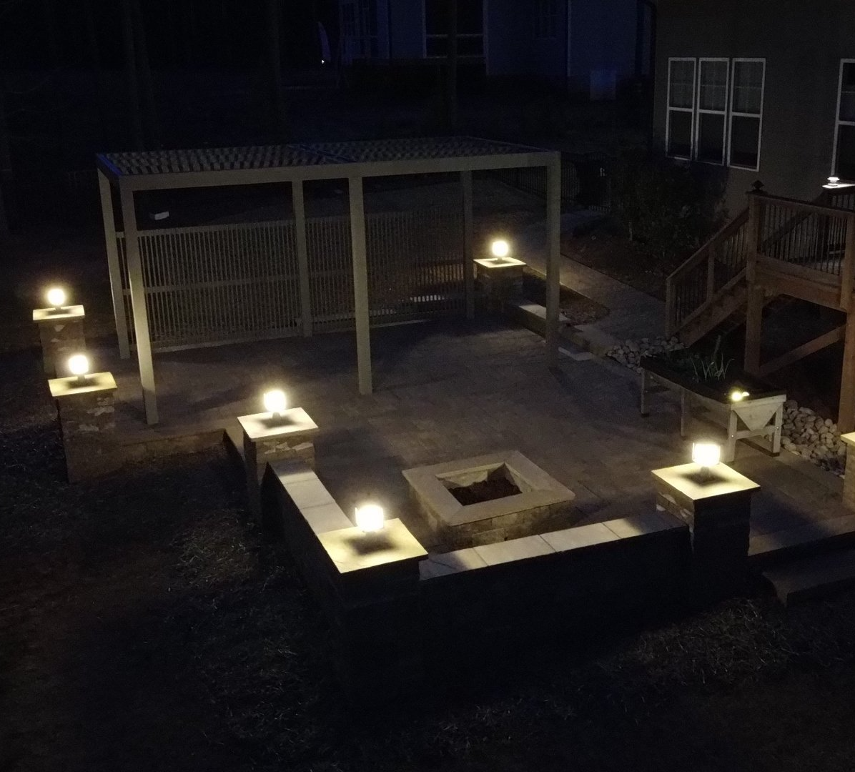 A stone patio at night features a square fire pit, a wooden pergola, and seating walls illuminated by warm post cap lights.