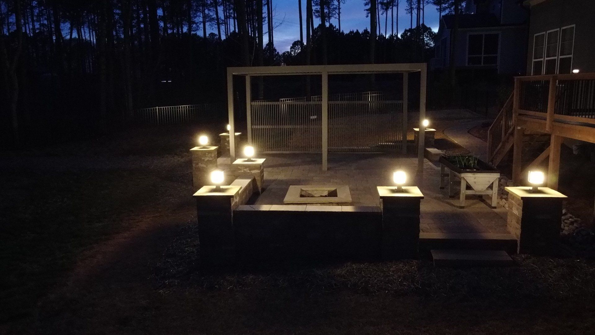An outdoor patio area at twilight featuring a central fire pit illuminated by glowing post-cap lights.