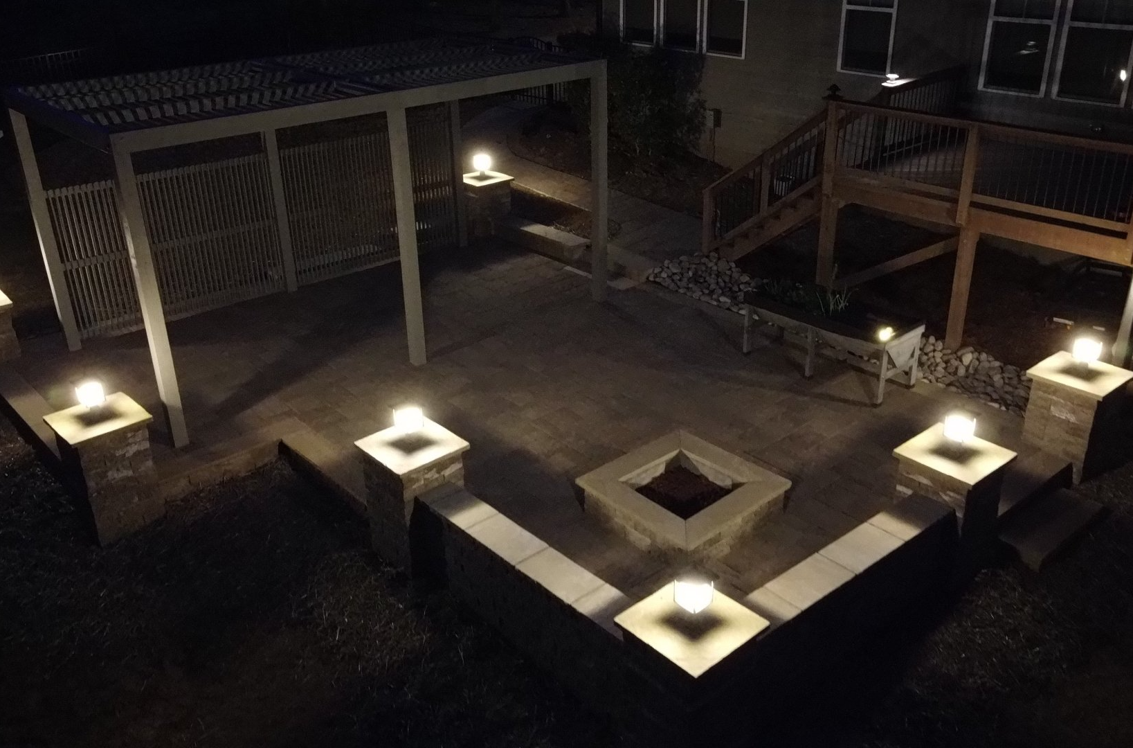 Nighttime outdoor patio with lit pillars, fire pit, pergola, and wooden deck.