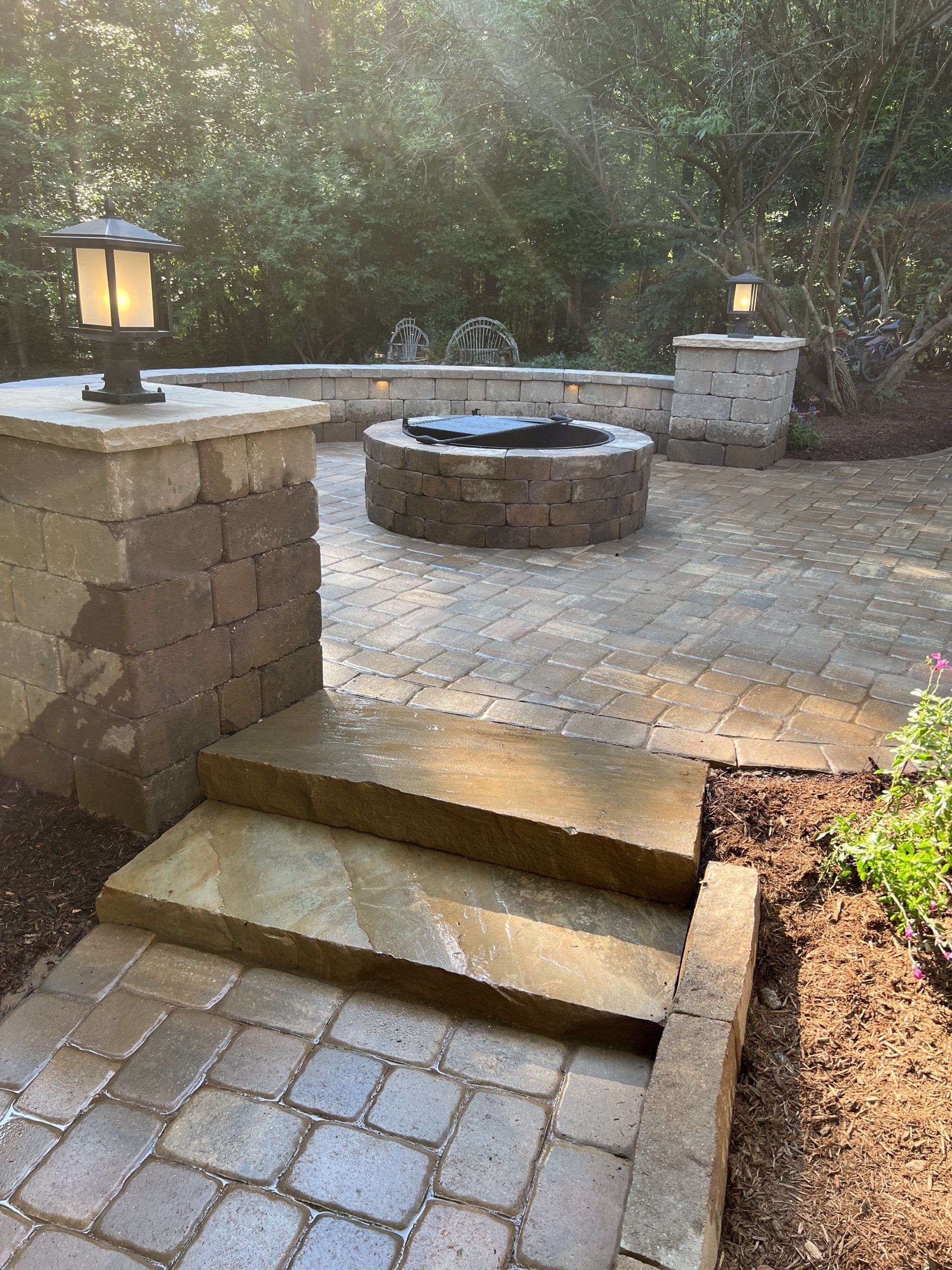 Brick patio with steps, fire pit, stone columns with lights, and greenery.