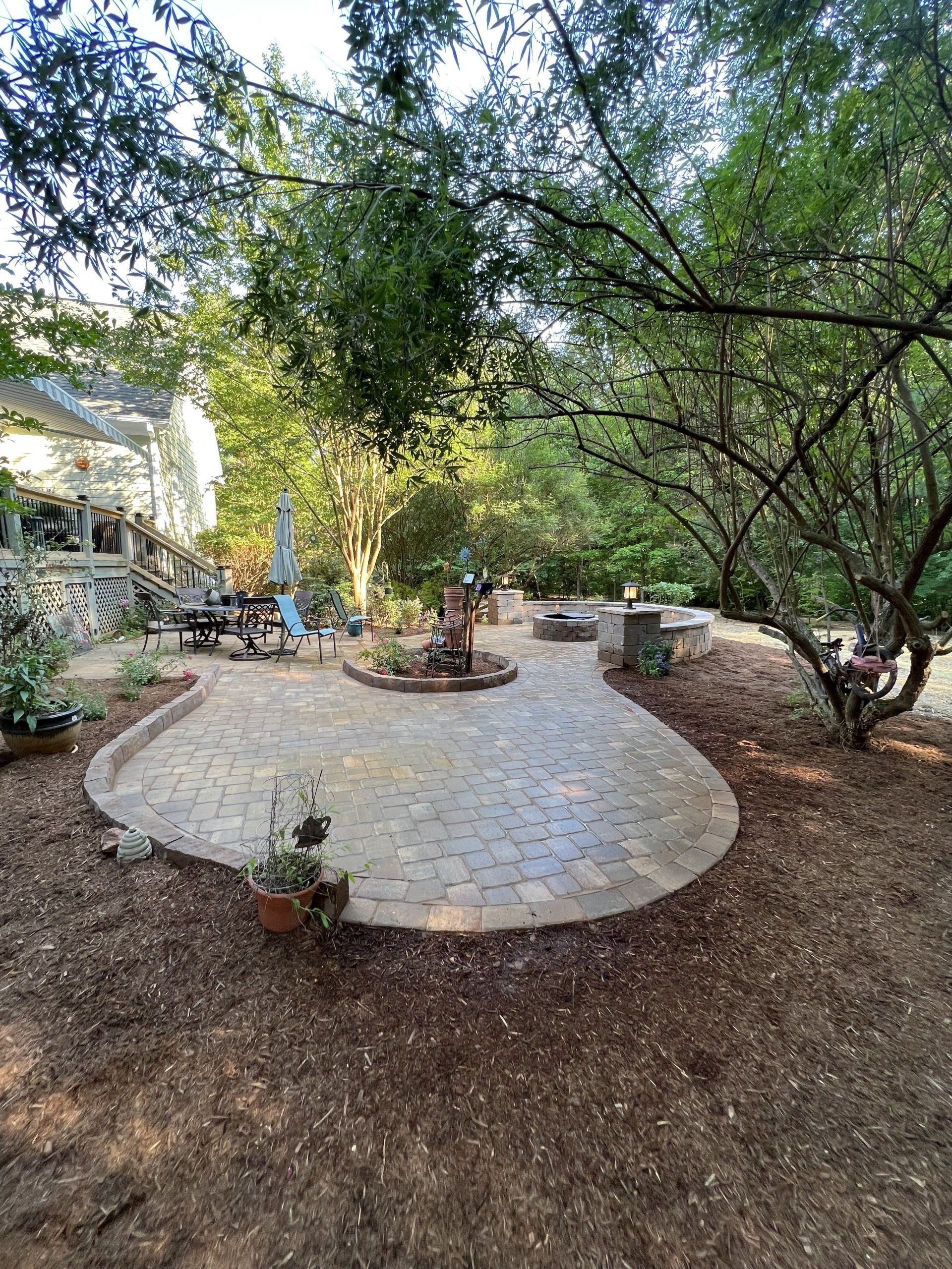 A landscaped backyard patio made of light-colored pavers, surrounded by trees, a mulch ground cover, and outdoor seating.