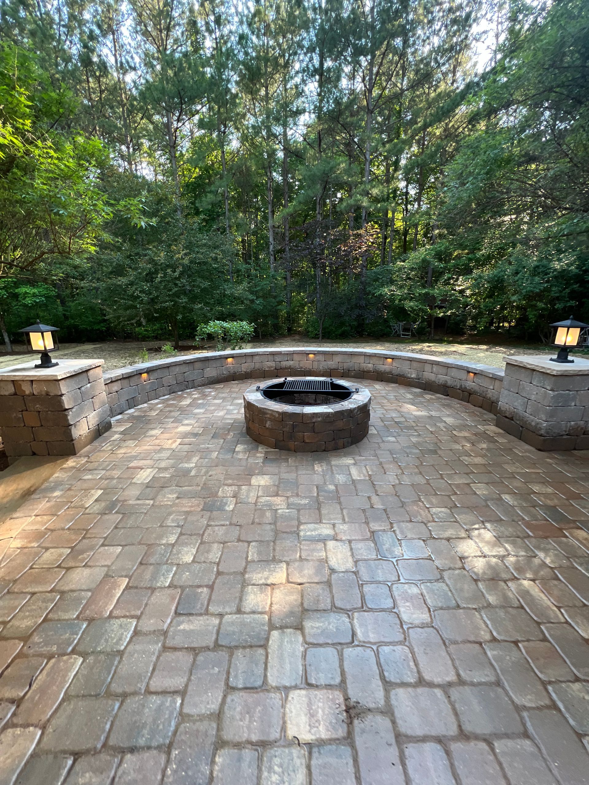 A circular stone fire pit sits on a paved patio bordered by a low, curved stone seating wall with two lamp posts.