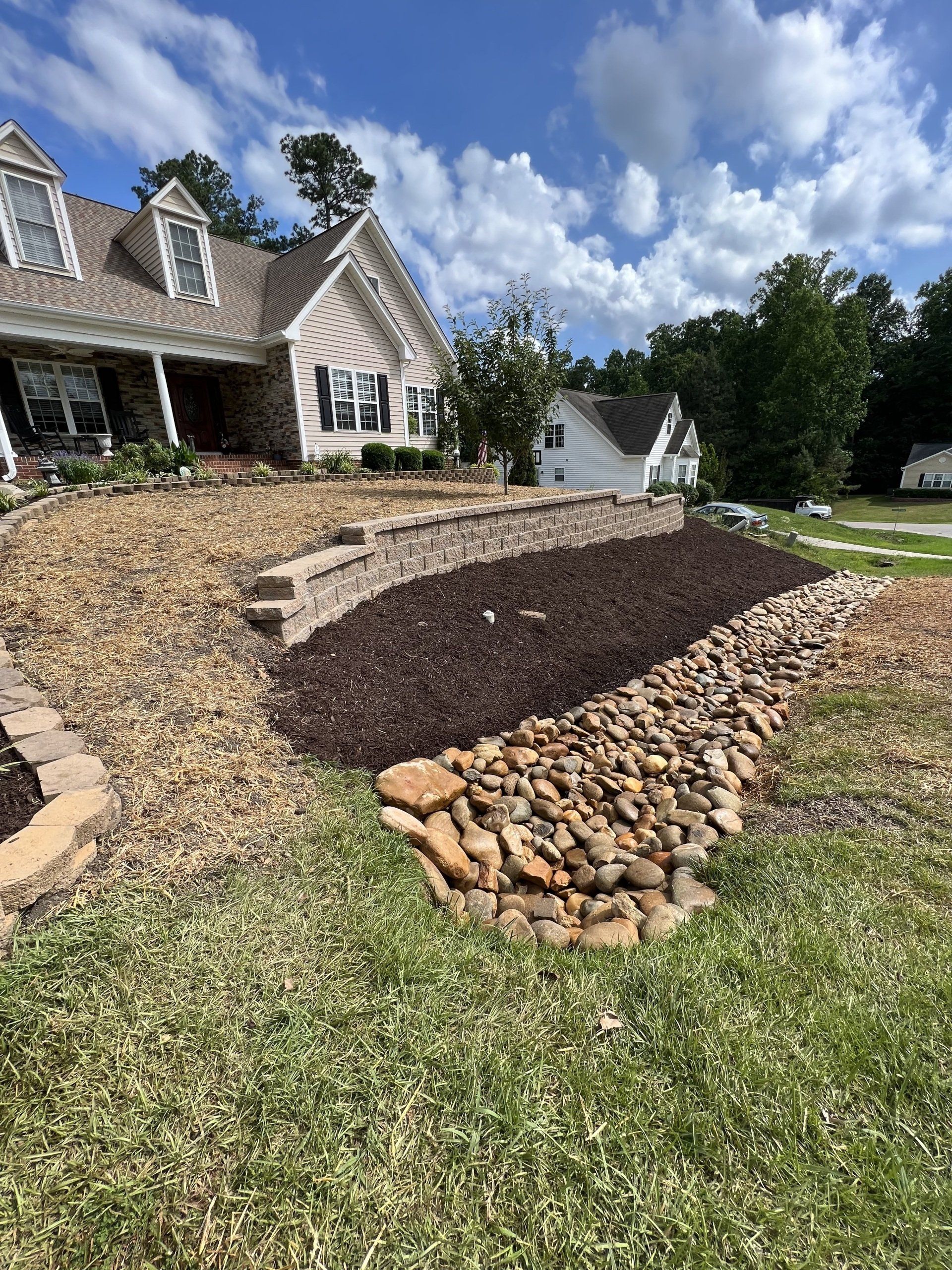 A suburban home features a tiered landscape with stone retaining walls, dark mulch beds, and a decorative rock drainage path.