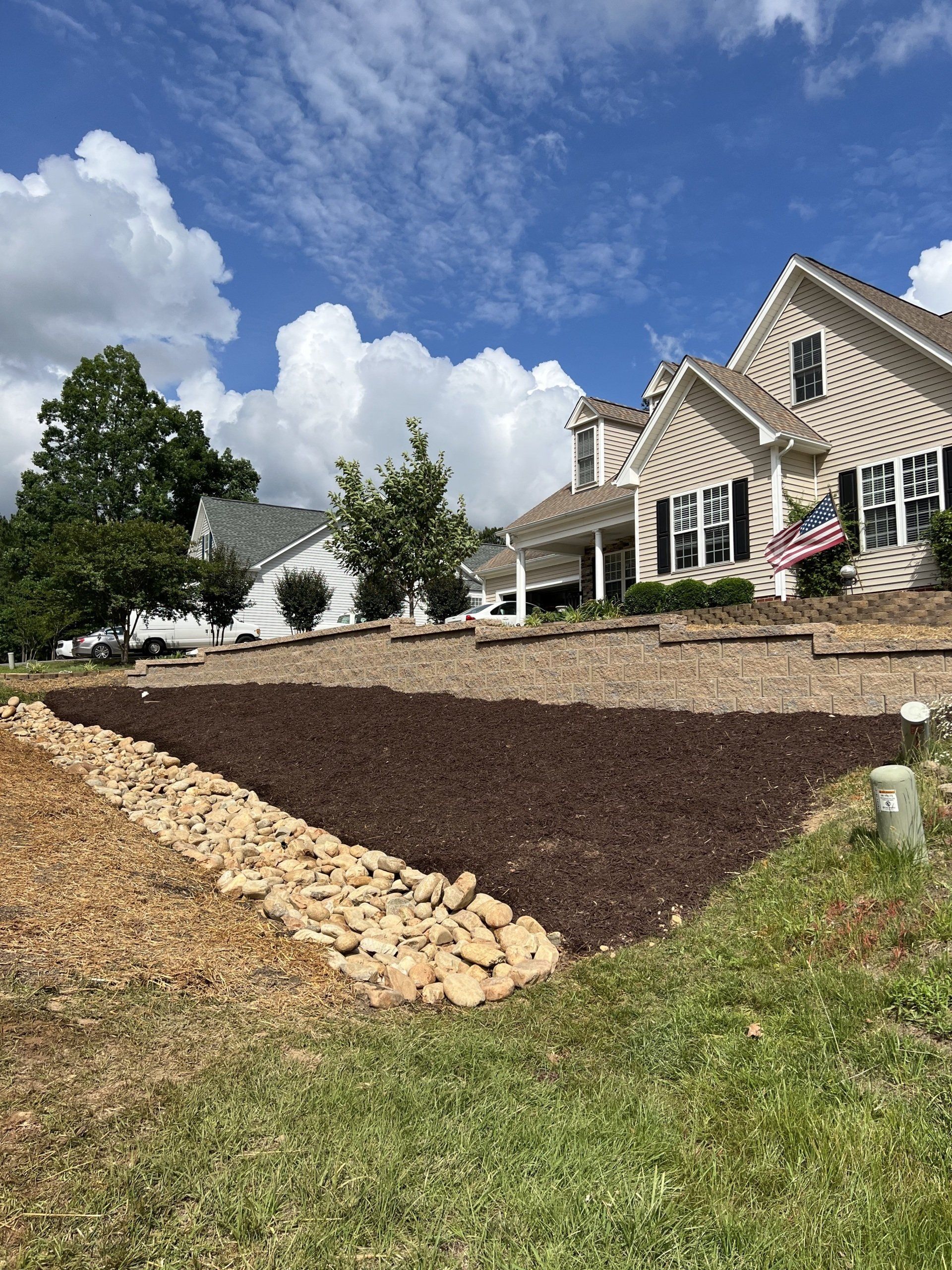 A residential front yard features landscaped terraces with dark mulch, stone borders, and a large house in the background.