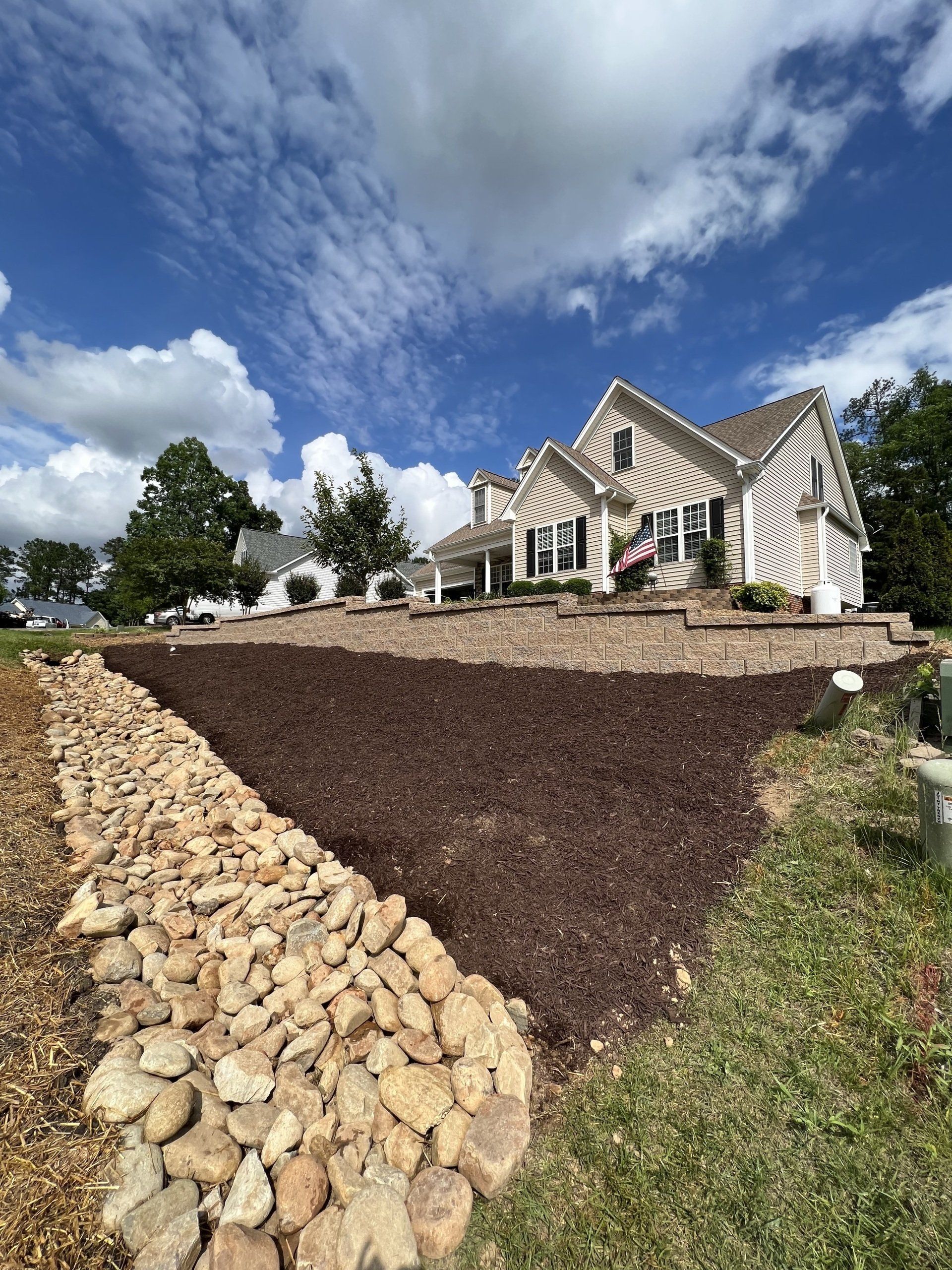 A large house with a steep roof sits behind a multi-tiered yard featuring a dark mulch slope and a large rock border.