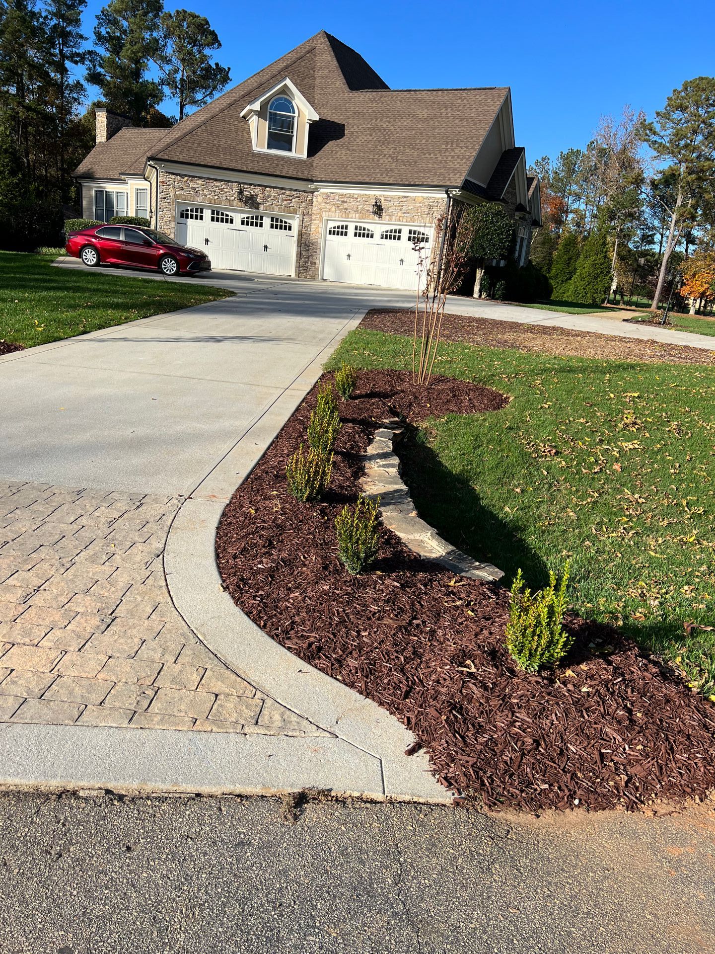 House with brick driveway, two-car garage, red car, and landscaped front yard with mulch and shrubs.