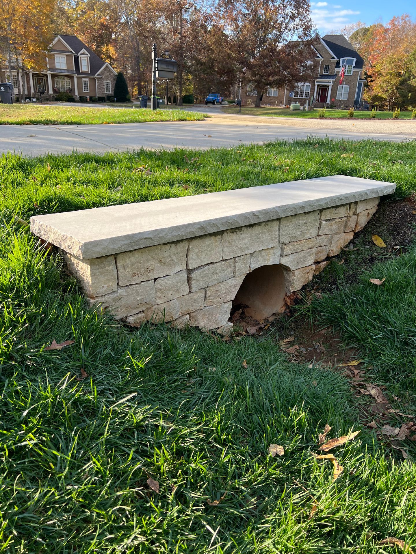 Stone-block bridge over a culvert in a grassy area, houses in the background, blue sky.