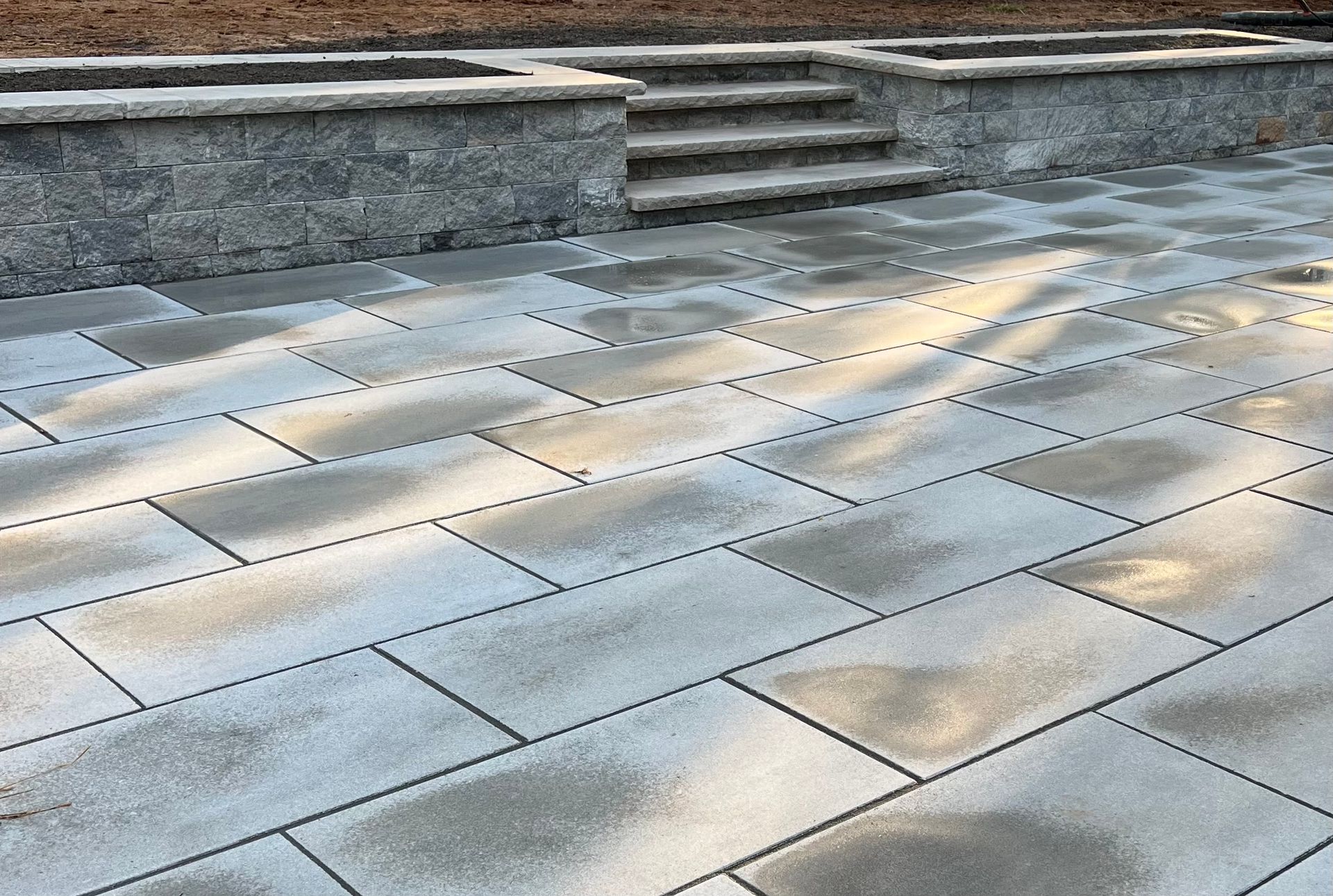 A light-colored stone patio with rectangular pavers and stone steps leading up to a matching low wall.
