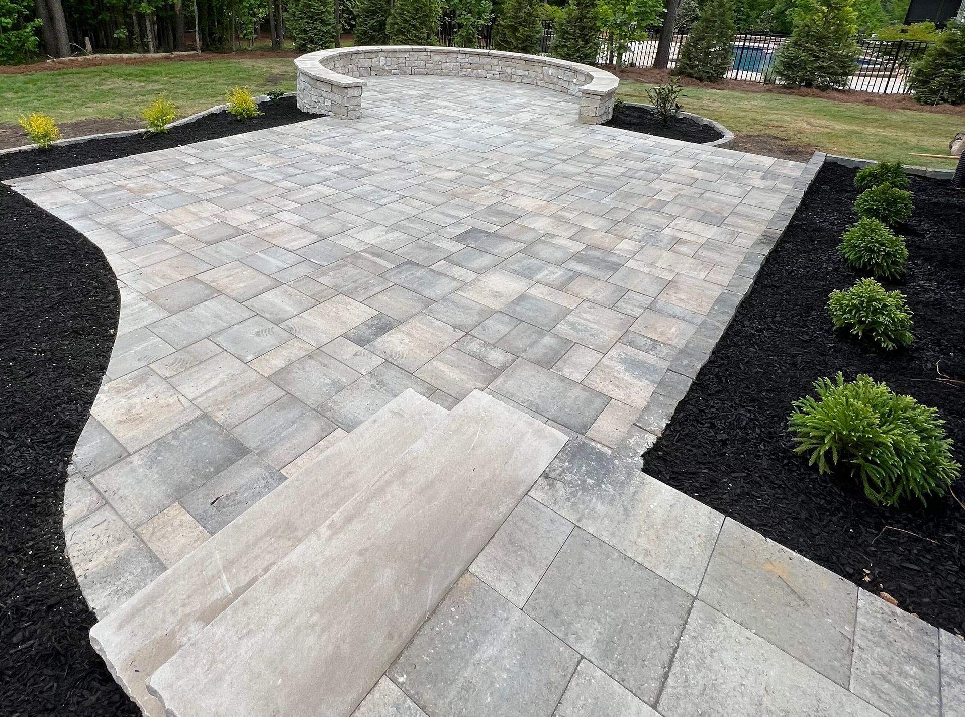 A stone paver patio with a curved stone retaining wall, surrounded by dark mulch and small shrubs in a grassy yard.