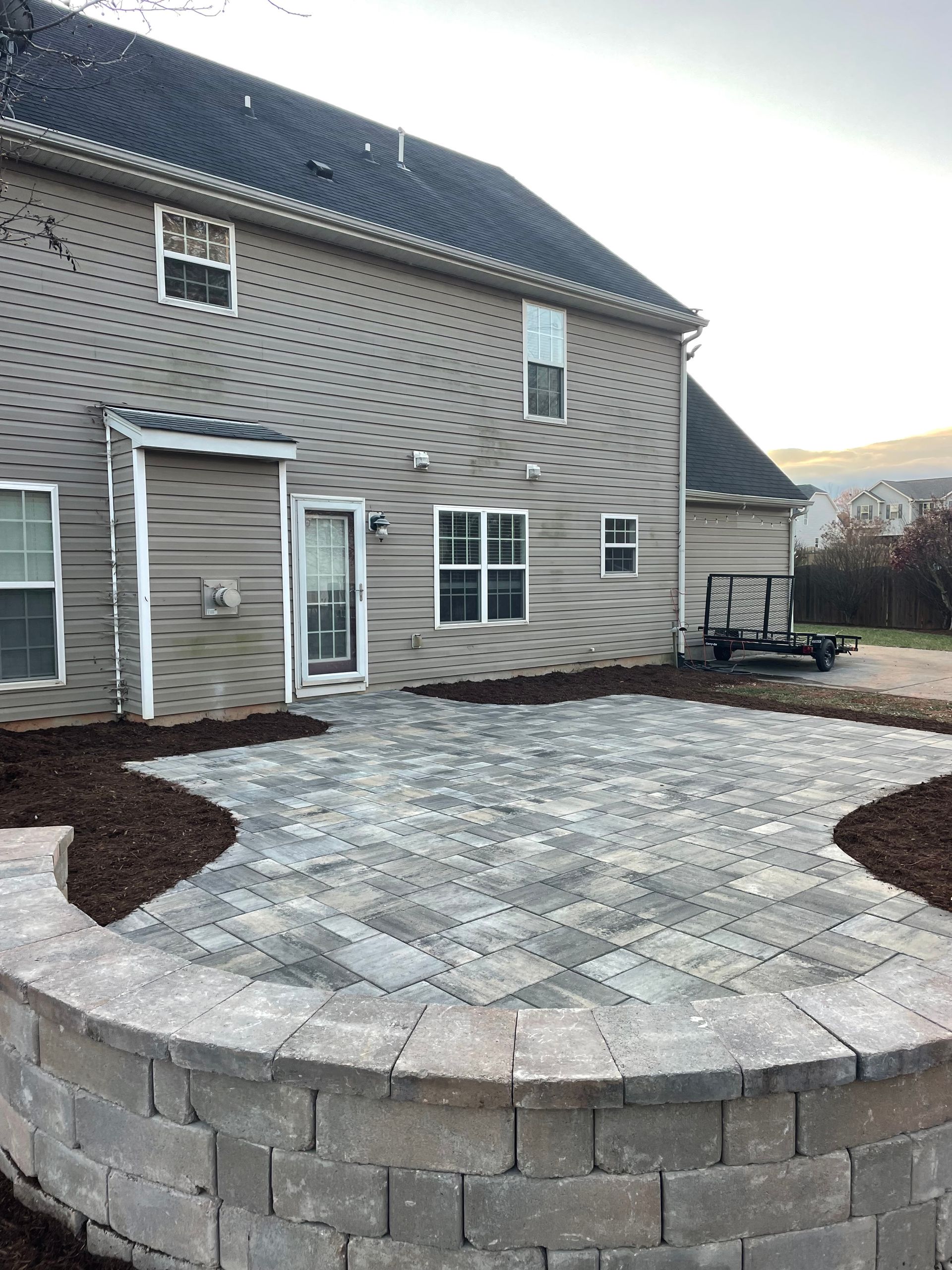 A newly installed stone patio and curved retaining wall in the backyard of a multi-story, tan-sided house.