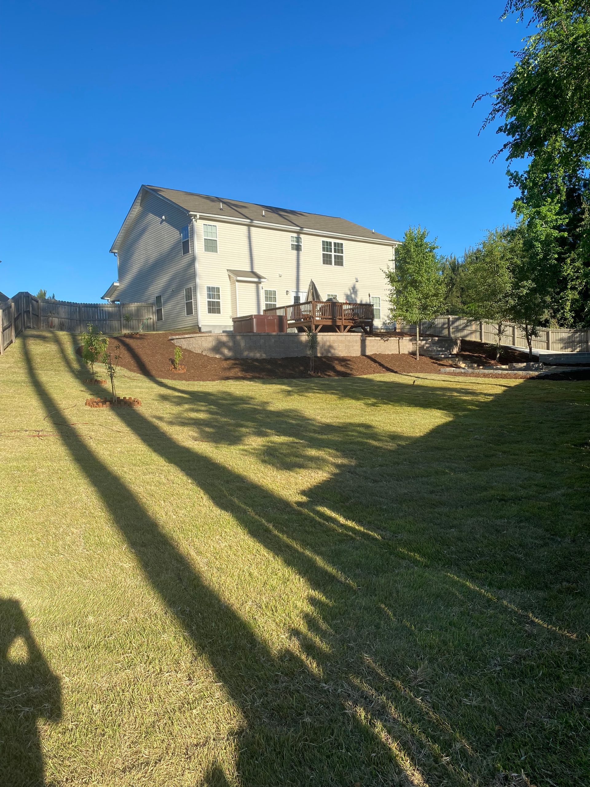 A two-story light-colored house with a backyard deck sits at the top of a grassy slope on a sunny day.