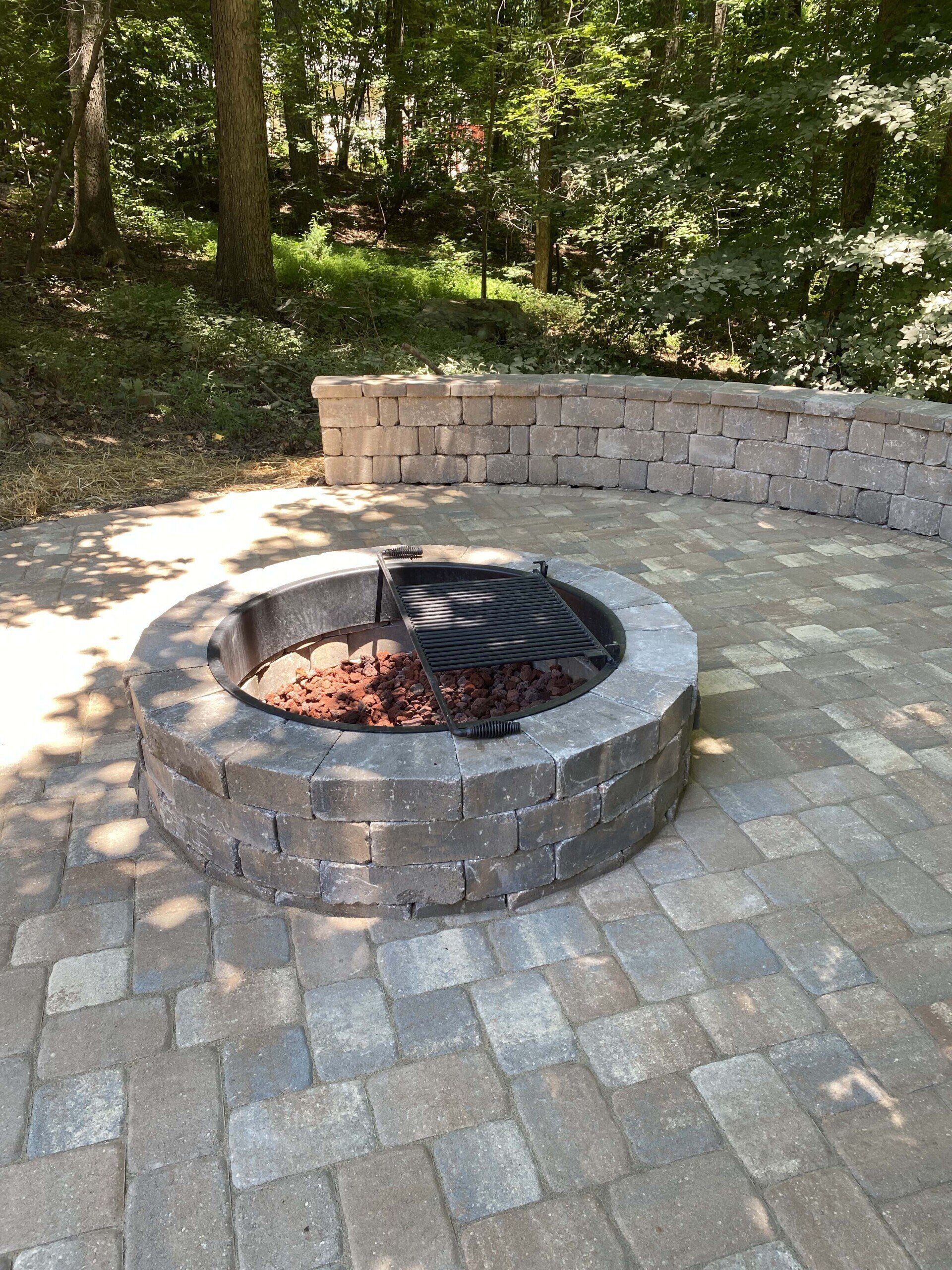 A circular stone fire pit with a metal cooking grate sits on a paver patio in front of a curved stone wall and forest.