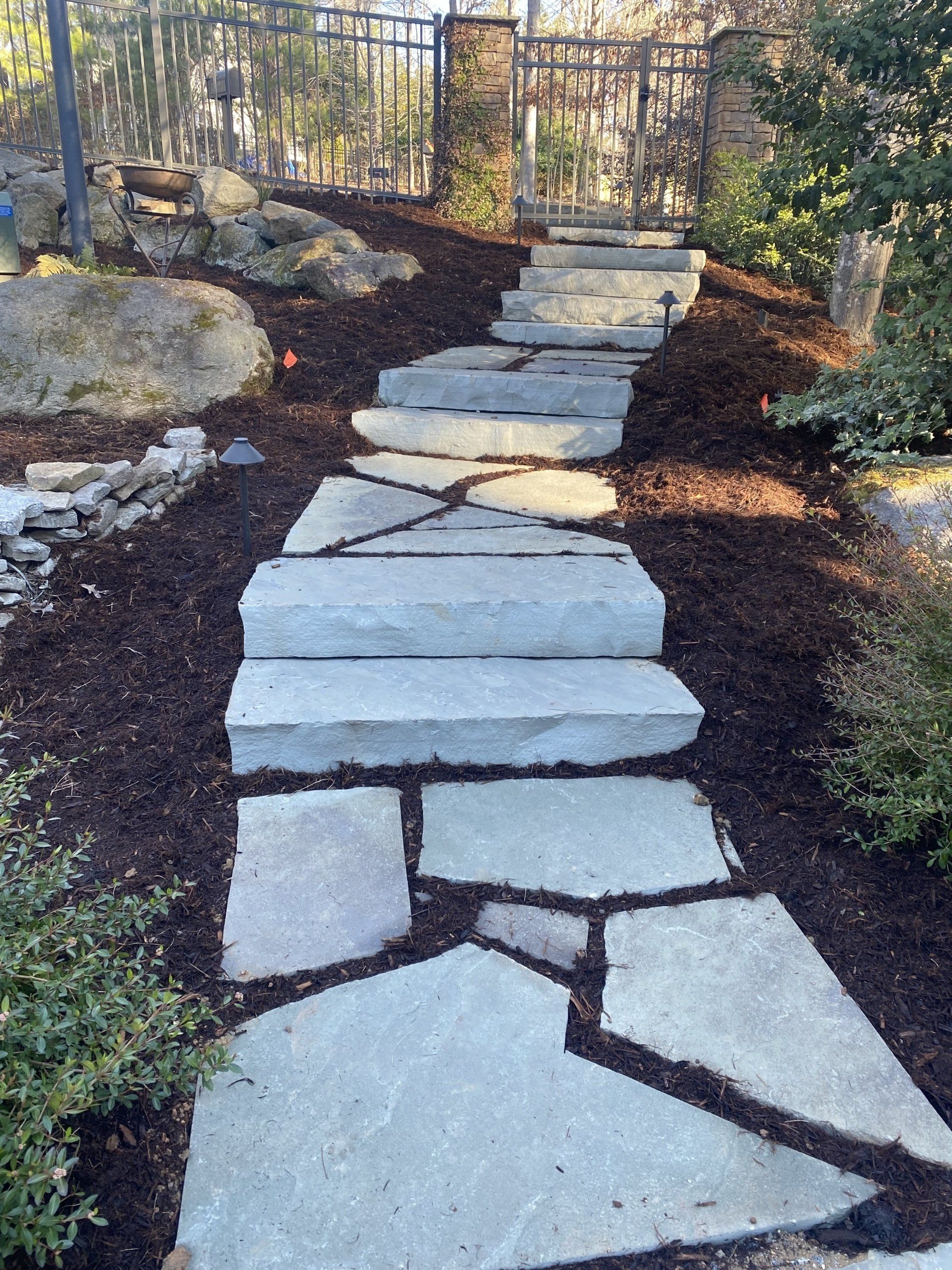 A stone pathway with wide, irregular steps leads uphill through dark mulch toward a gate in a fenced backyard.